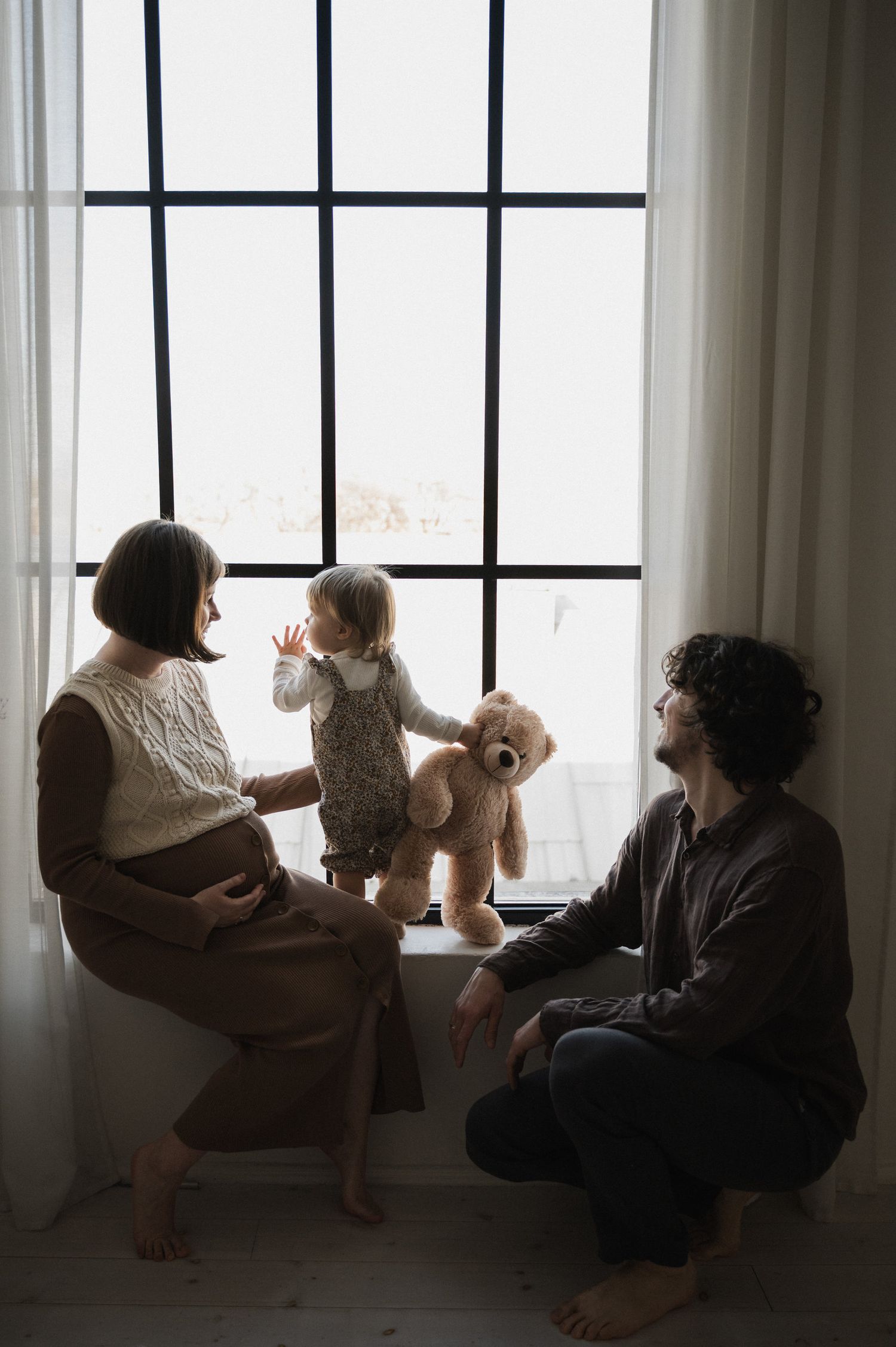 Family sitting by large windows in a bright white room with natural light streaming in.