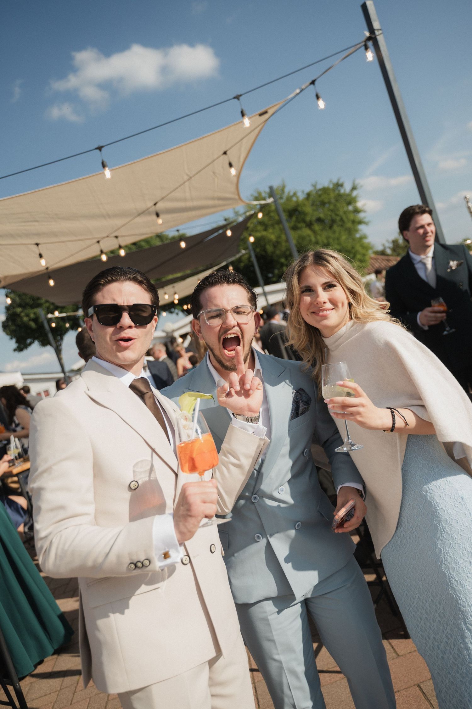 Three elegantly dressed wedding guests smiling and holding cocktails at an outdoor reception venue.