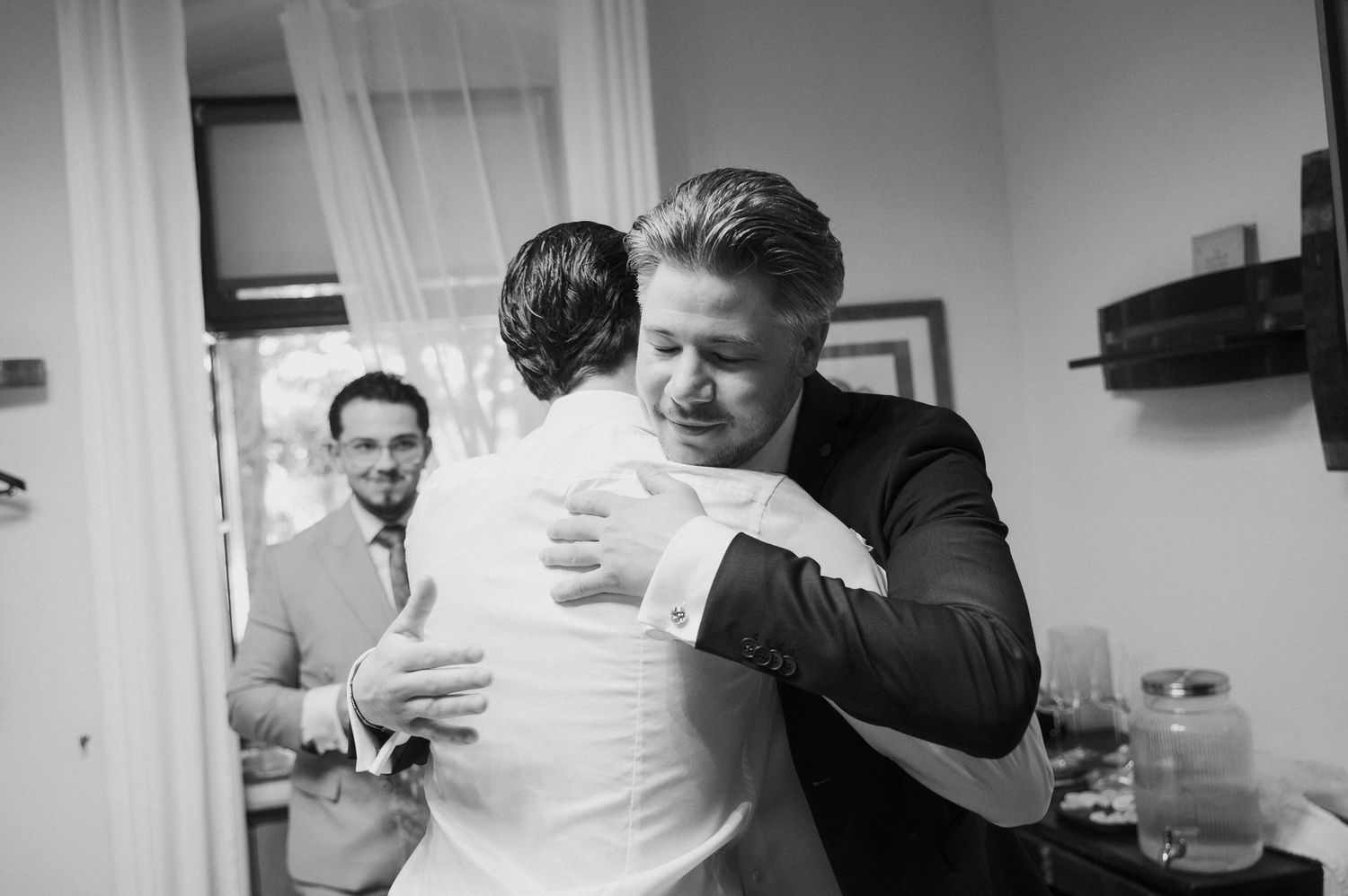 Black and white photo of two men sharing an emotional hug at a wedding, with a guest smiling in the background.