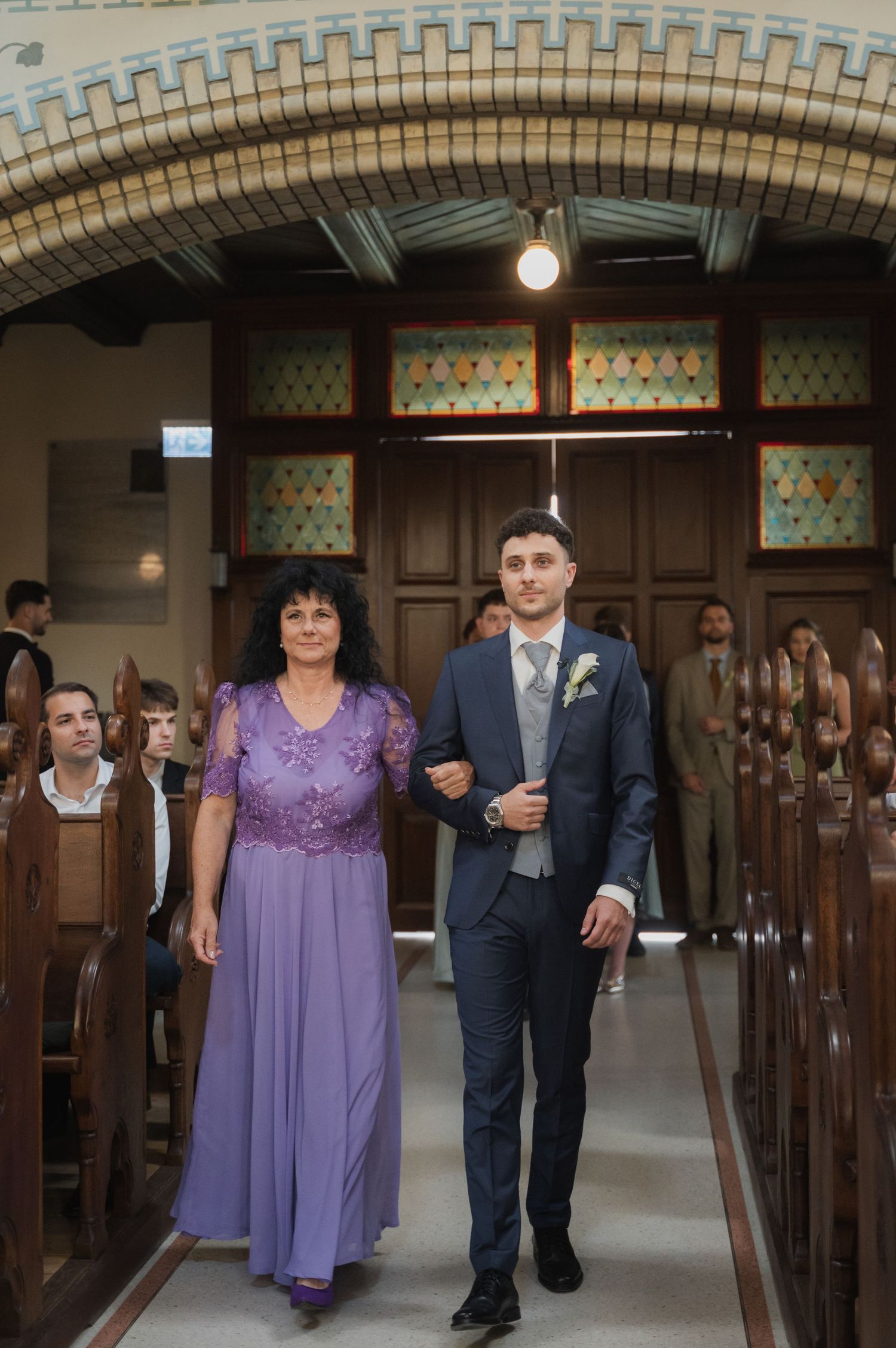 Groom in navy suit walks down church aisle with woman in purple dress during wedding ceremony.