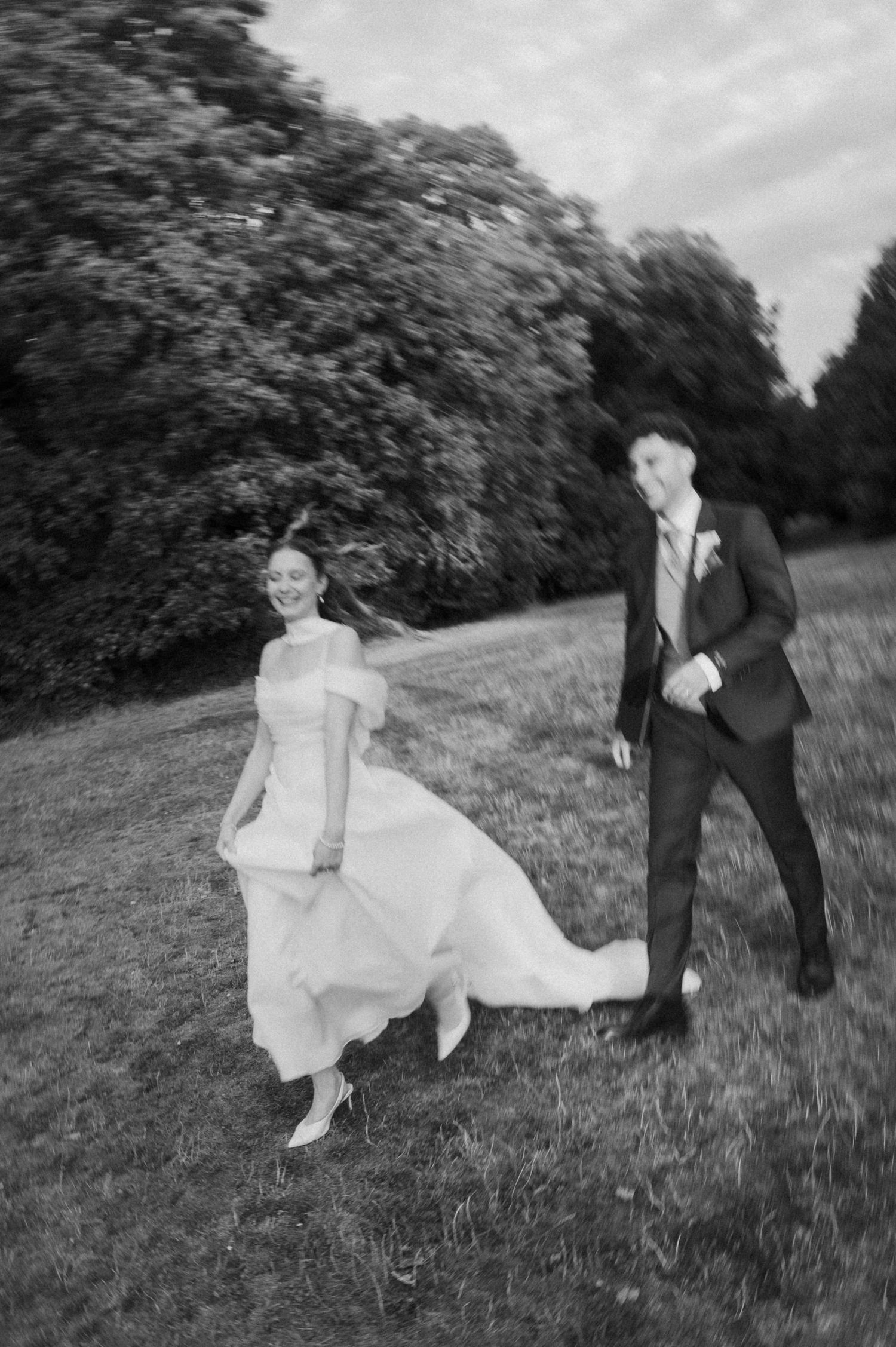 Black and white photo of a bride and groom joyfully running across a lush green lawn on their wedding day.