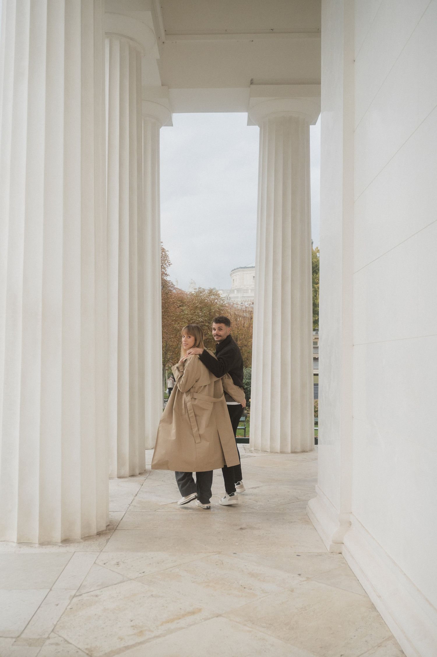 People walking through a modern colonnade with white pillars and stone floor in an architectural setting.