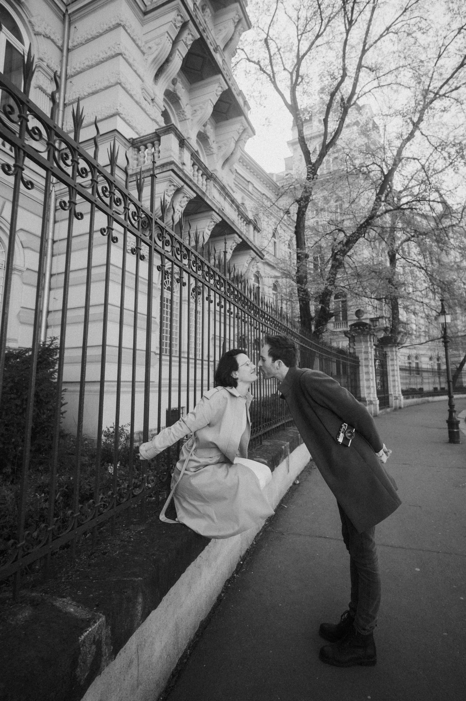 A romantic black and white street scene with two people leaning towards each other by an ornate iron fence in a city.
