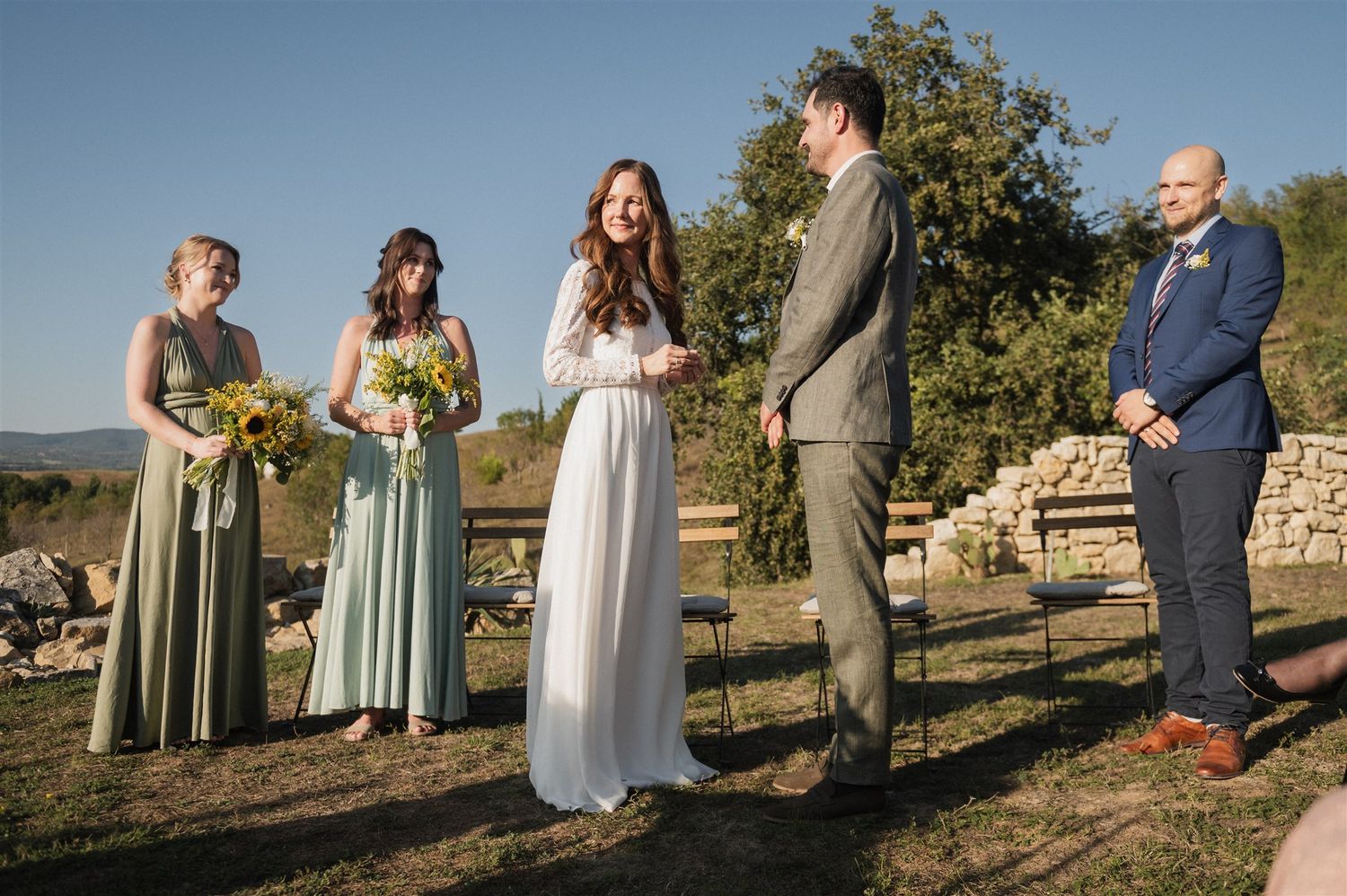 Bridesmaids in sage green dresses and guests gather for an outdoor wedding ceremony on a hillside.