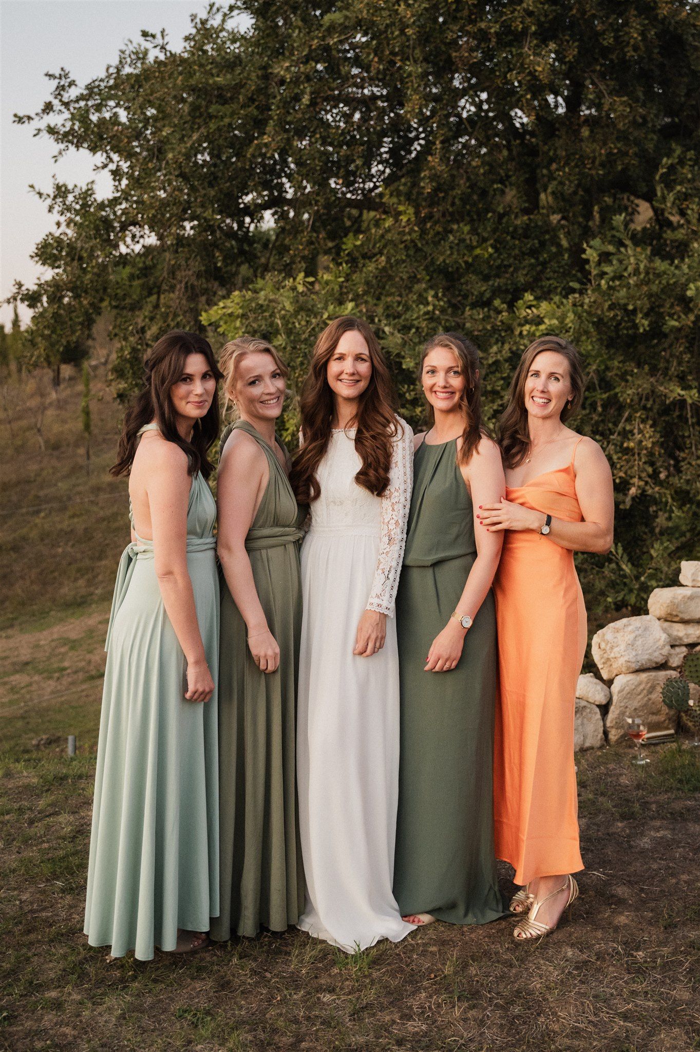 A group in coordinating dresses in shades of mint green, olive, white and peach pose together outdoors.