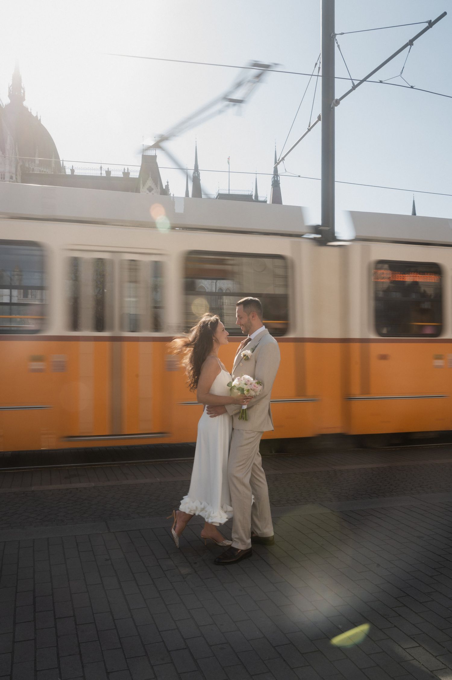 A couple embraces romantically as an orange tram passes by in the background.