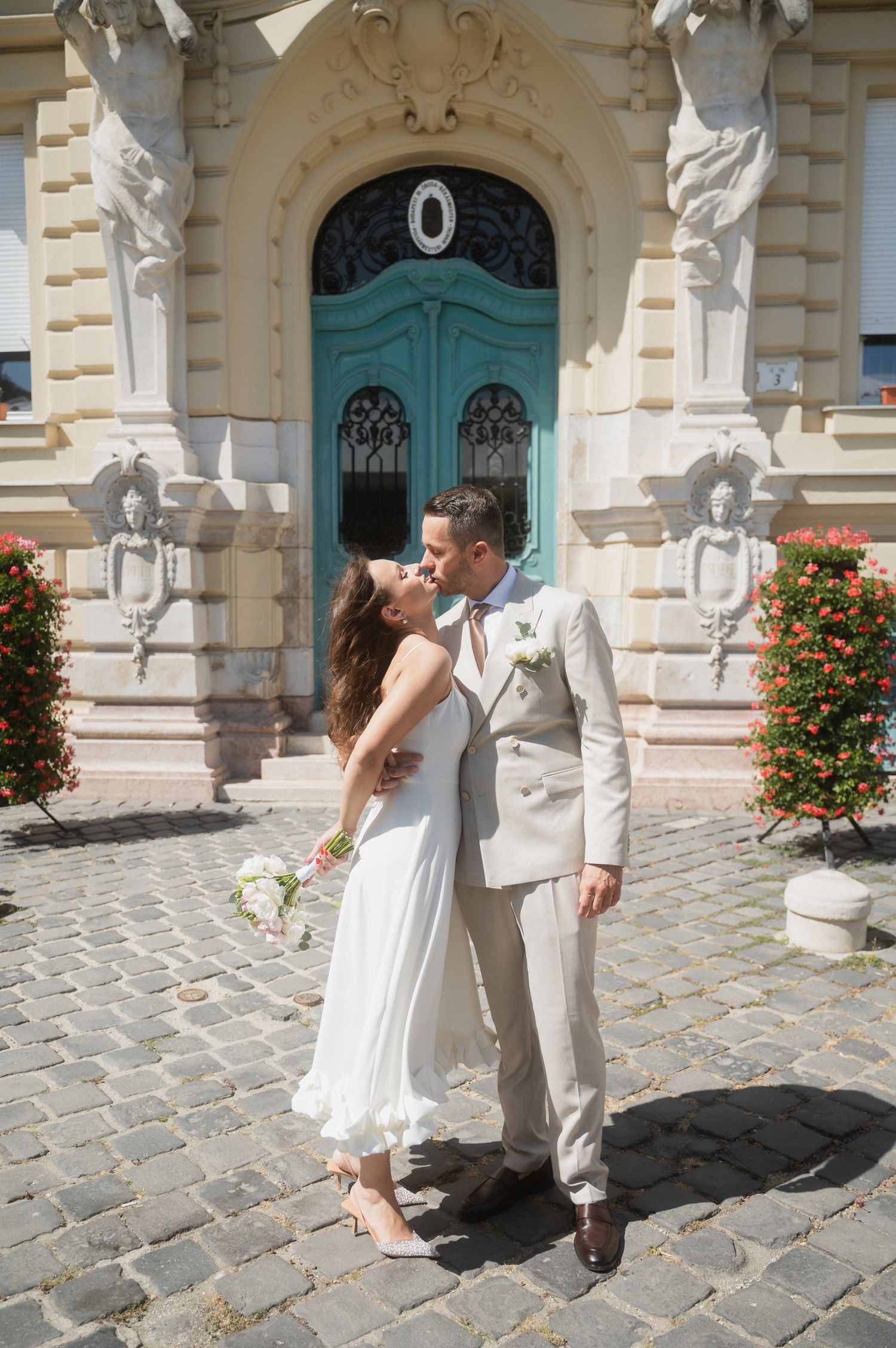 A couple in wedding attire dances playfully on cobblestone streets in front of an ornate building with a turquoise door.