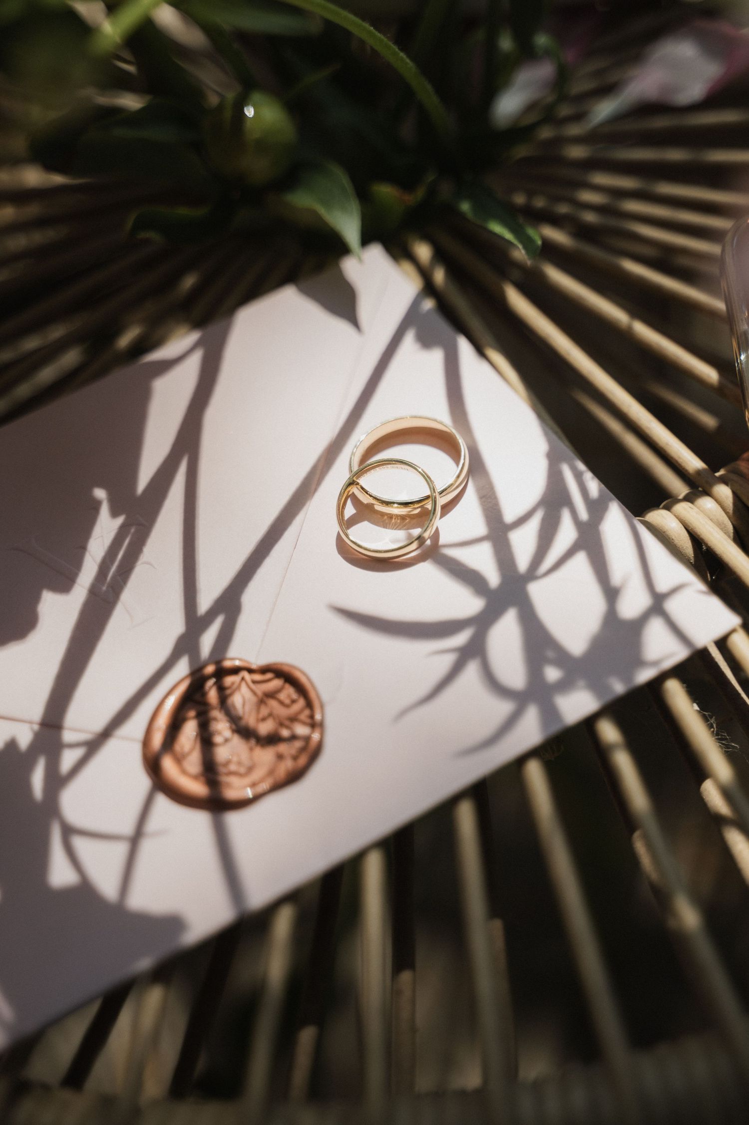 Gold wedding rings resting on a white envelope with wax seal, surrounded by bamboo and tropical leaf shadows.