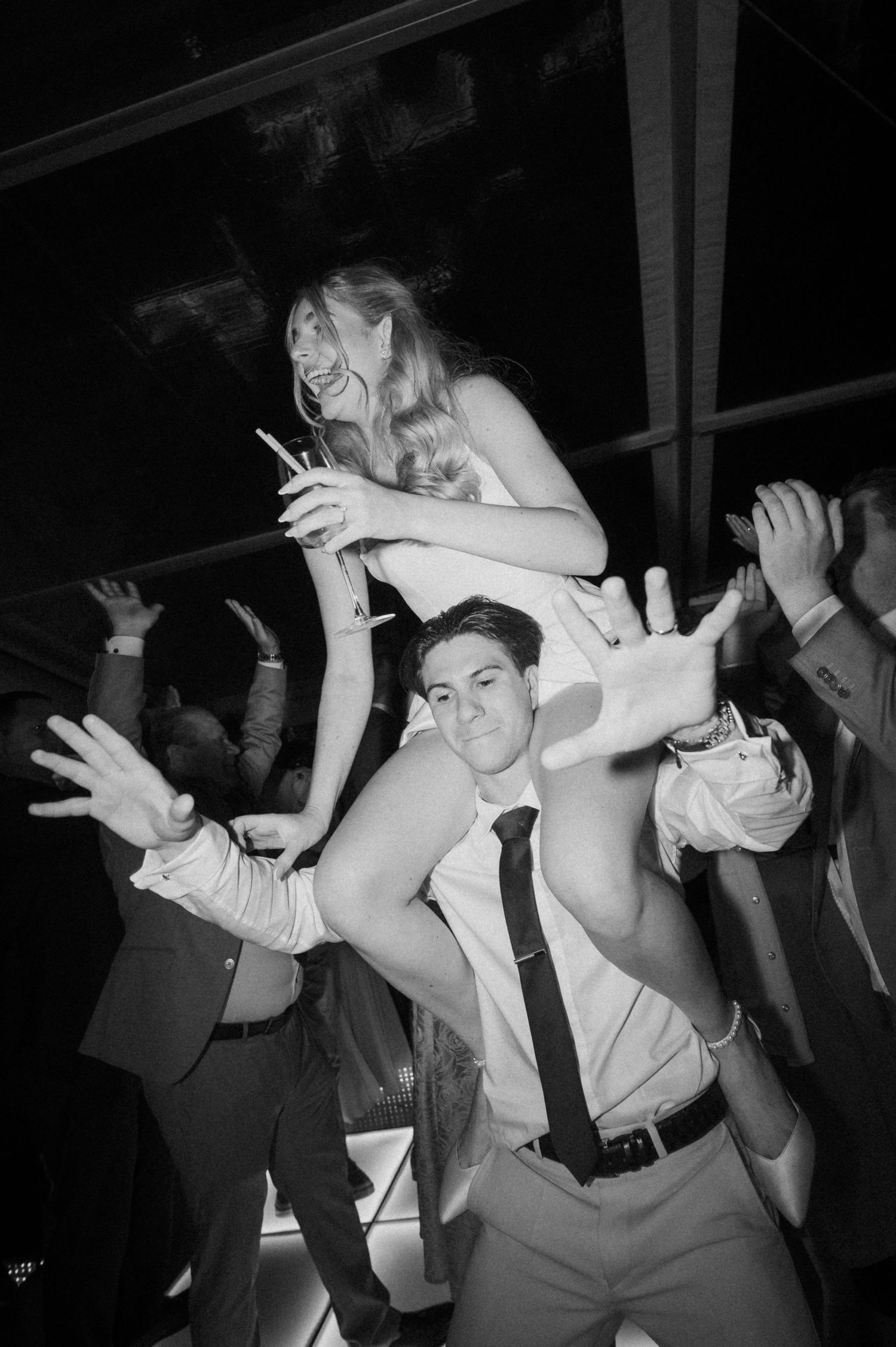 Black and white photo of a woman sitting on a man's shoulders dancing at a lively wedding reception party.