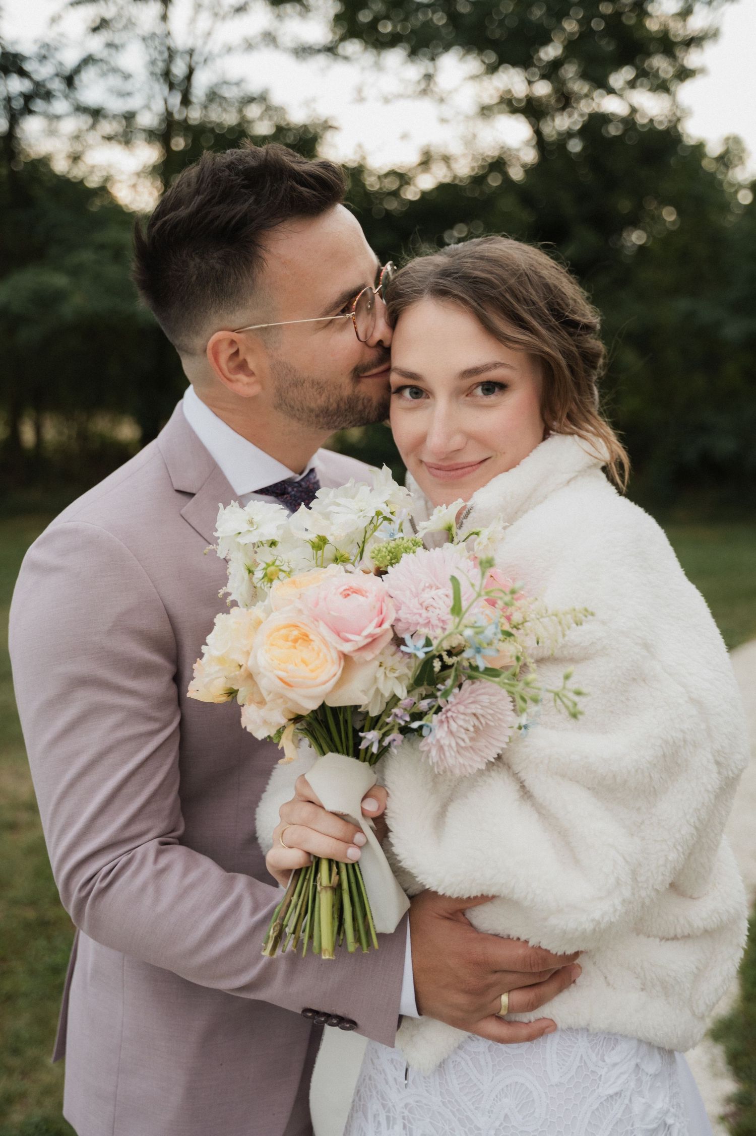 Groom in pink suit kisses bride on cheek as she smiles holding pastel bouquet outdoors.