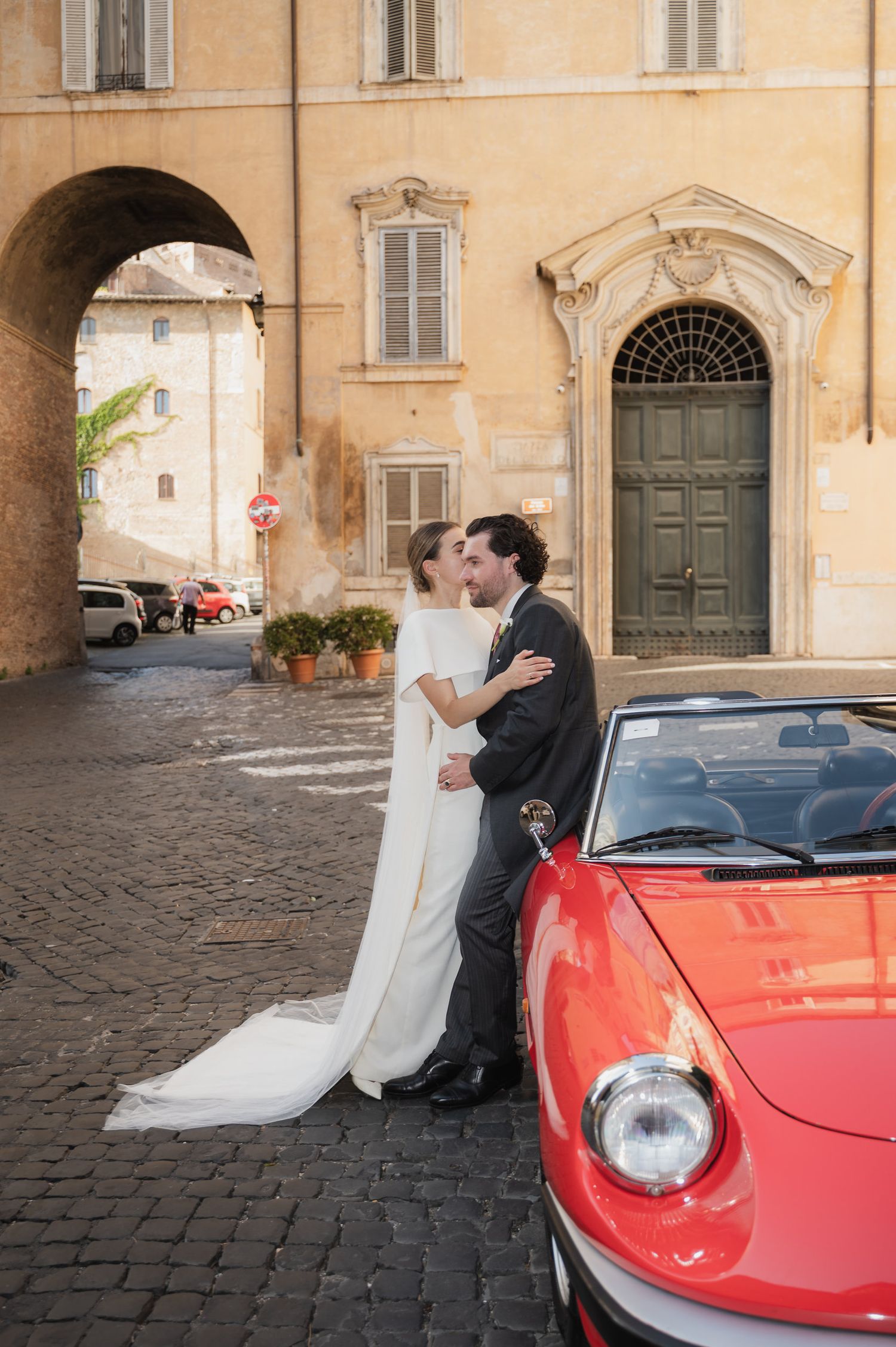 A tender wedding moment against warm Italian architecture with a red sports car.