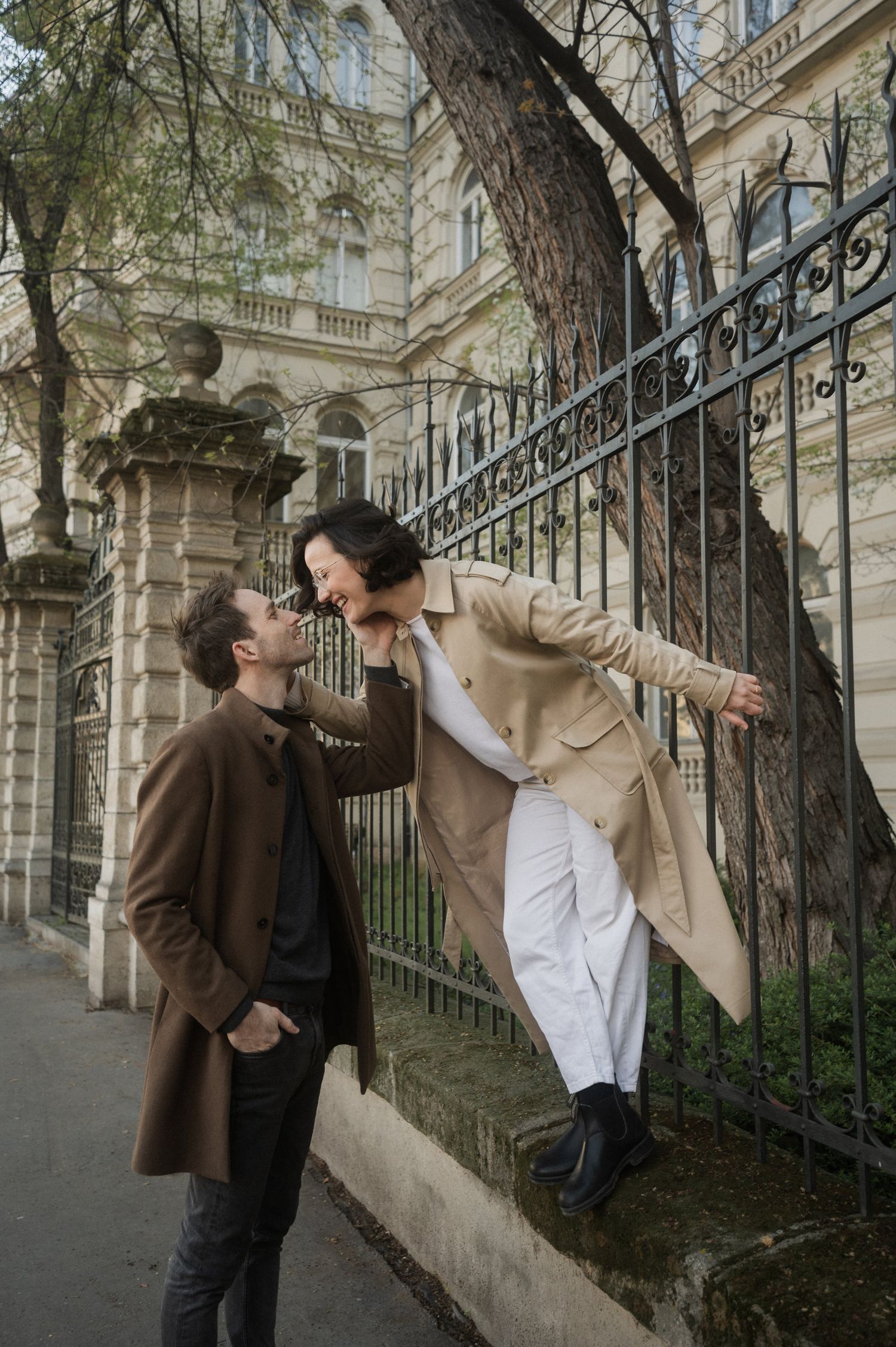 Two people playfully interact in a dramatic sequence along an ornate iron fence and historic building exterior.