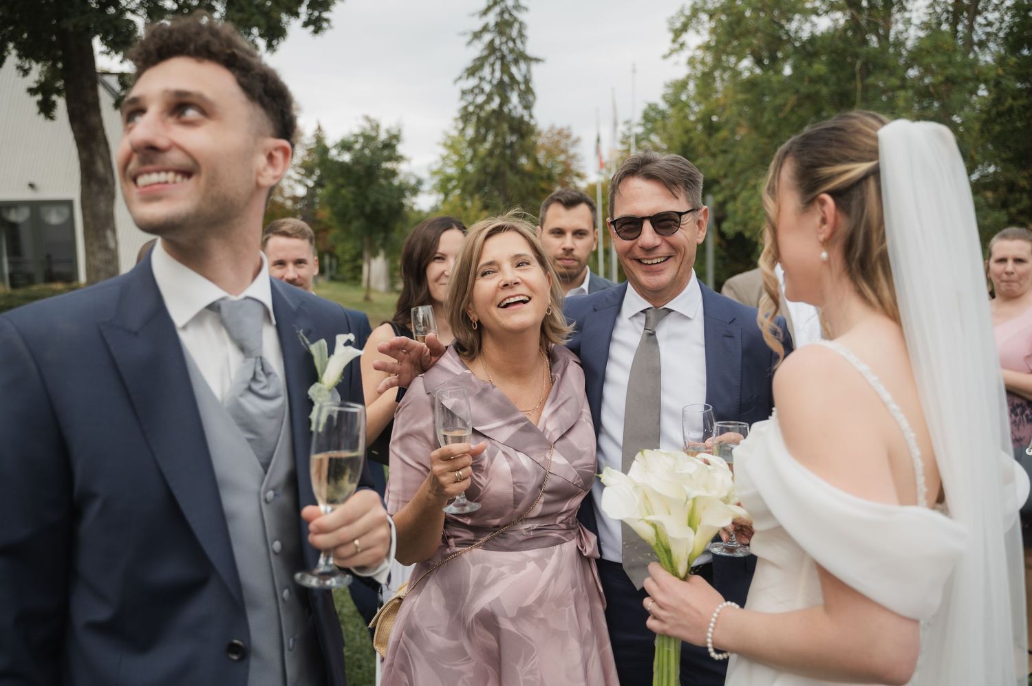 Joyful wedding guests celebrate outdoors with champagne as bride in white veil smiles with bouquet nearby.