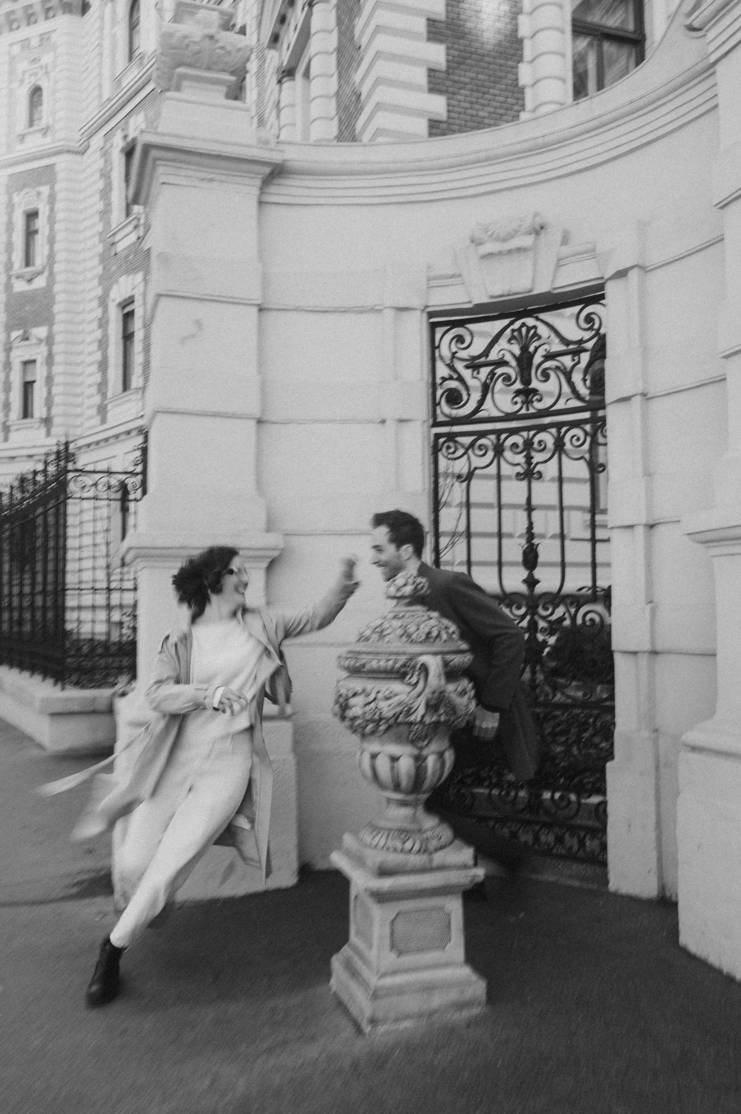A vintage black and white photo shows a playful scene between two people near an ornate stone entrance with iron gates.