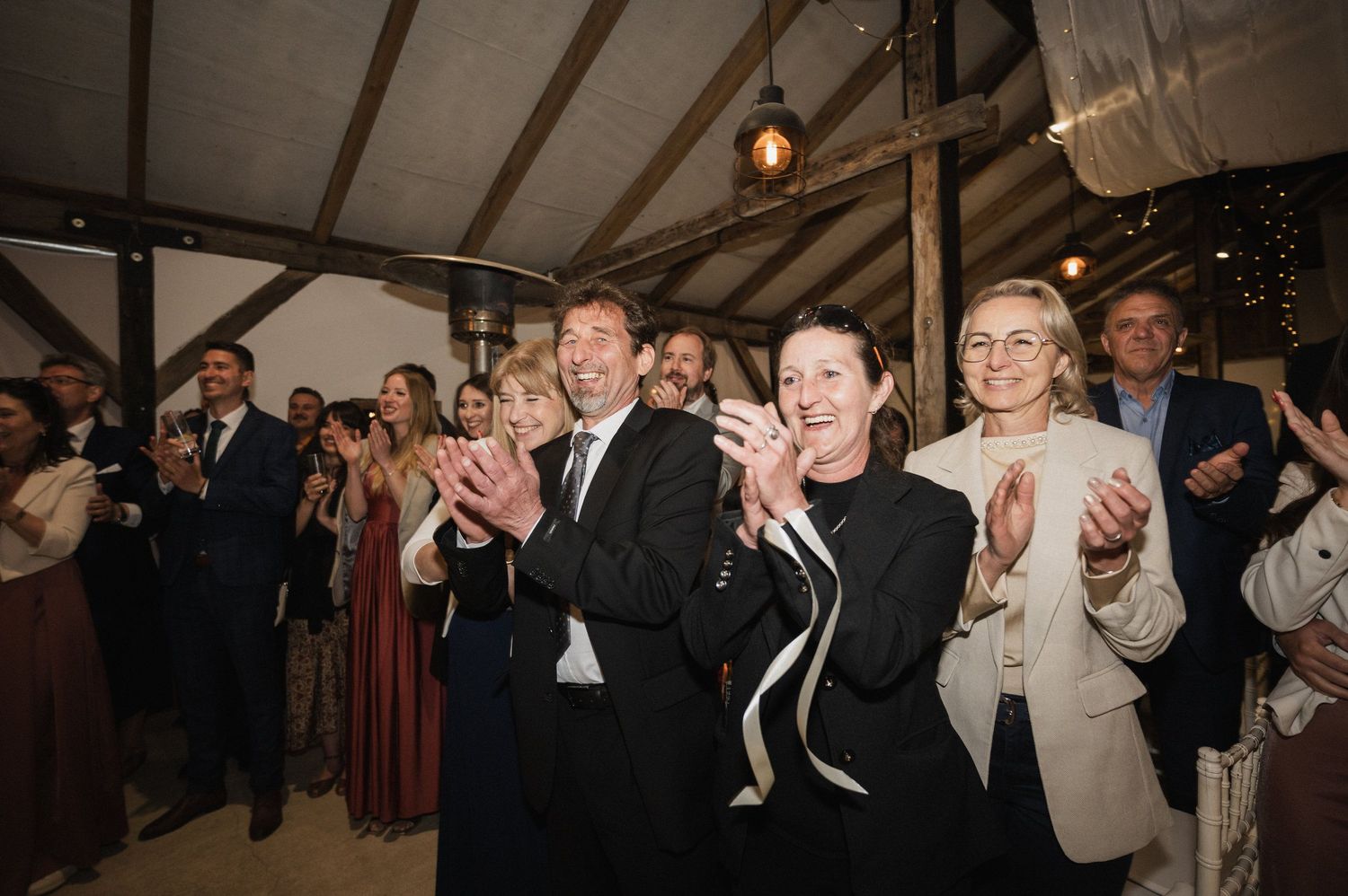 Wedding guests clap and cheer during a celebration in a rustic tent venue with string lights overhead.