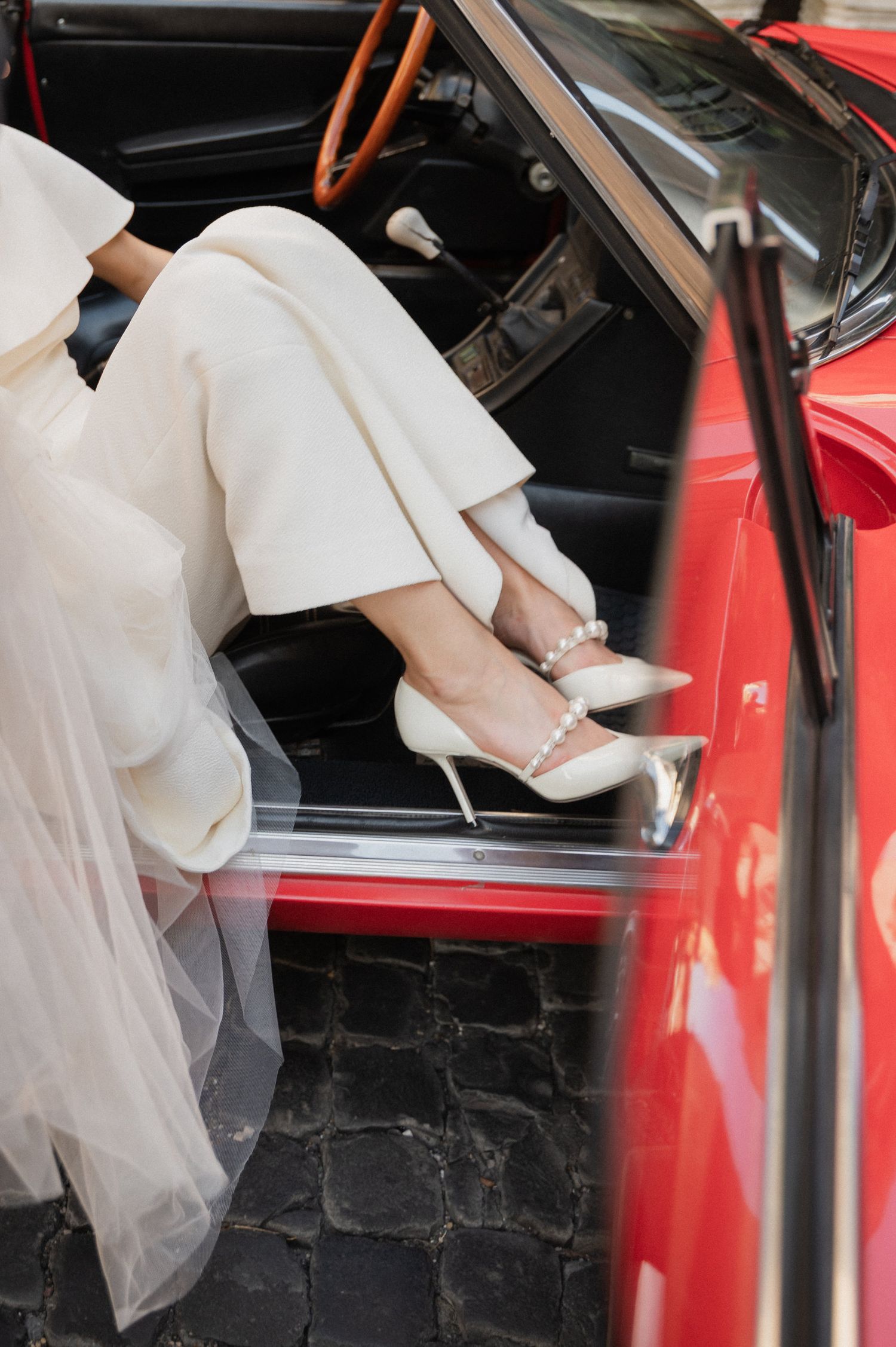 Close-up of white wedding dress and veil as bride enters classic red car.