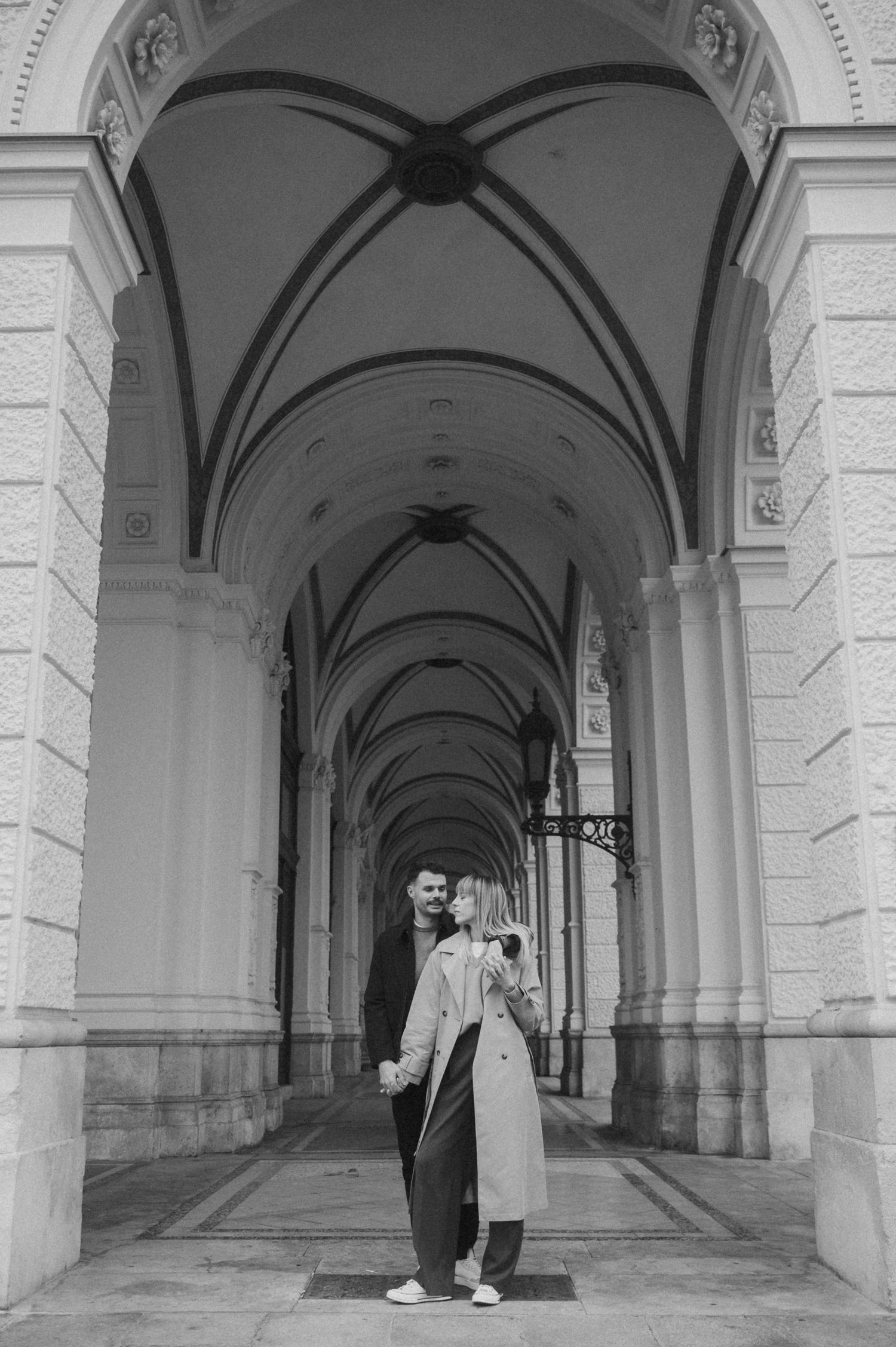 People stand in a historic stone archway corridor with vaulted ceilings and repeating architectural columns.