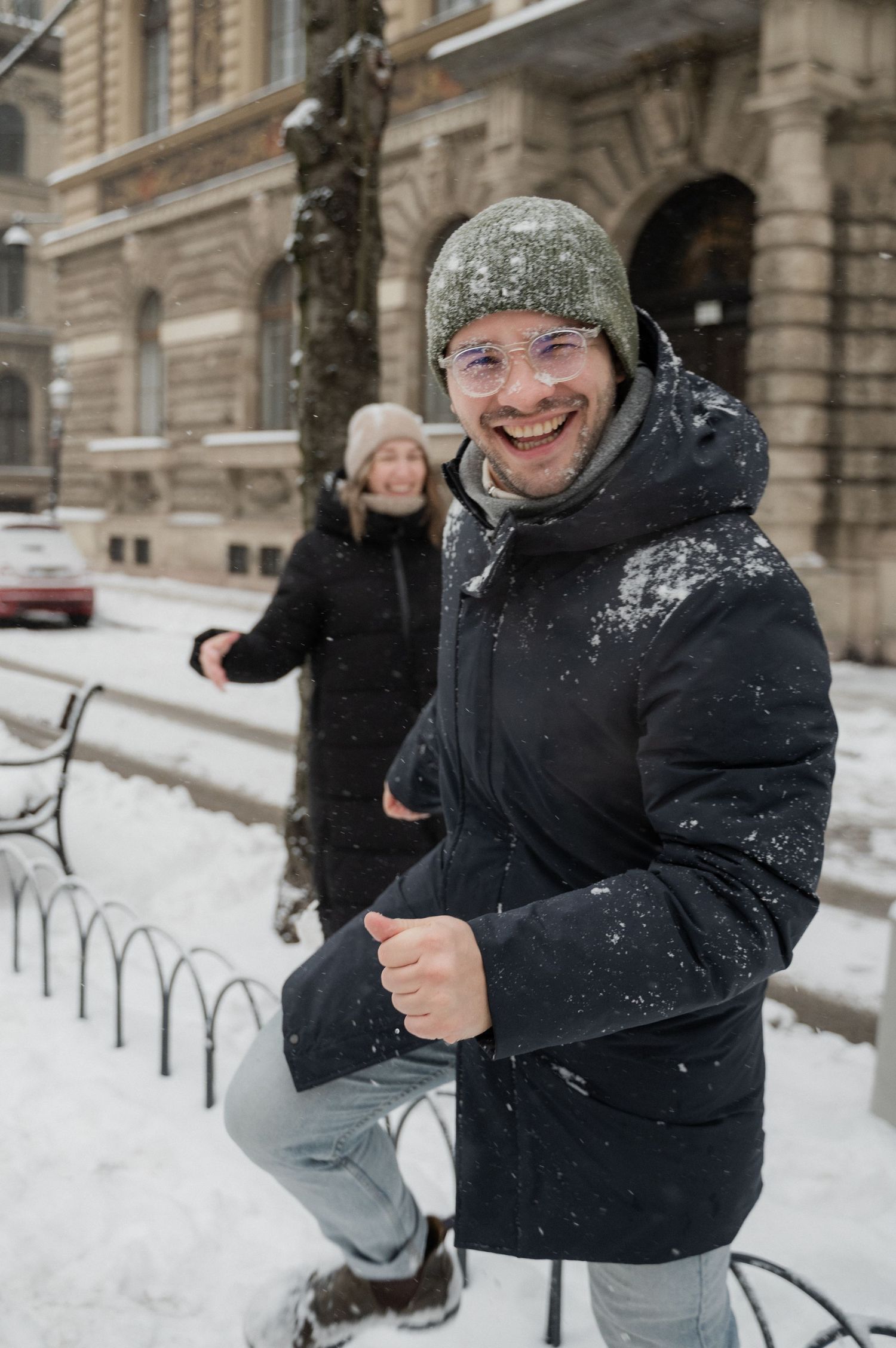 A person in winter gear smiling joyfully in front of a snow-covered historic building.