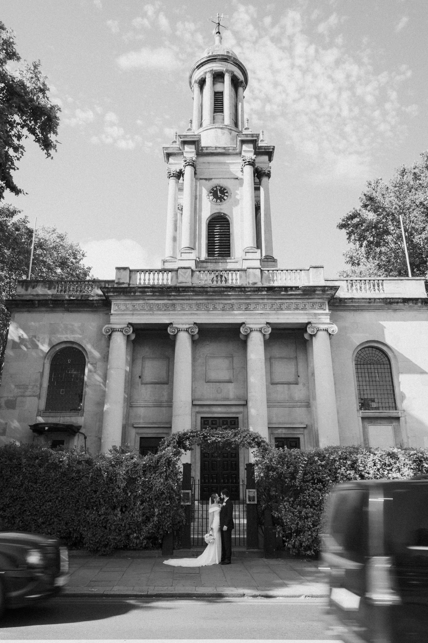 Black and white photo of a bride and groom standing before a grand neoclassical church with a clock tower in London.