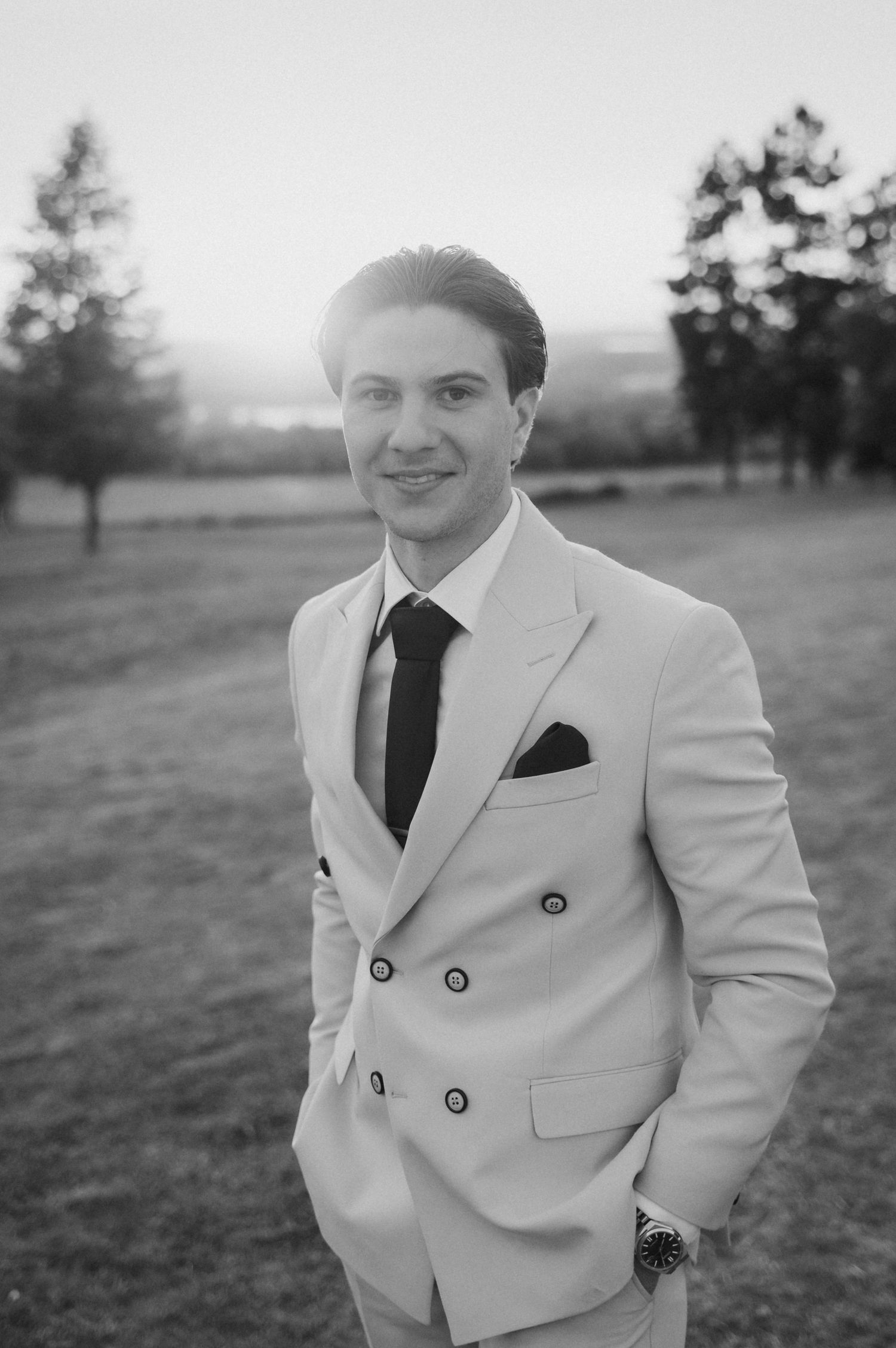 Elegant groom in double-breasted white suit with black tie smiling outdoors in black and white.