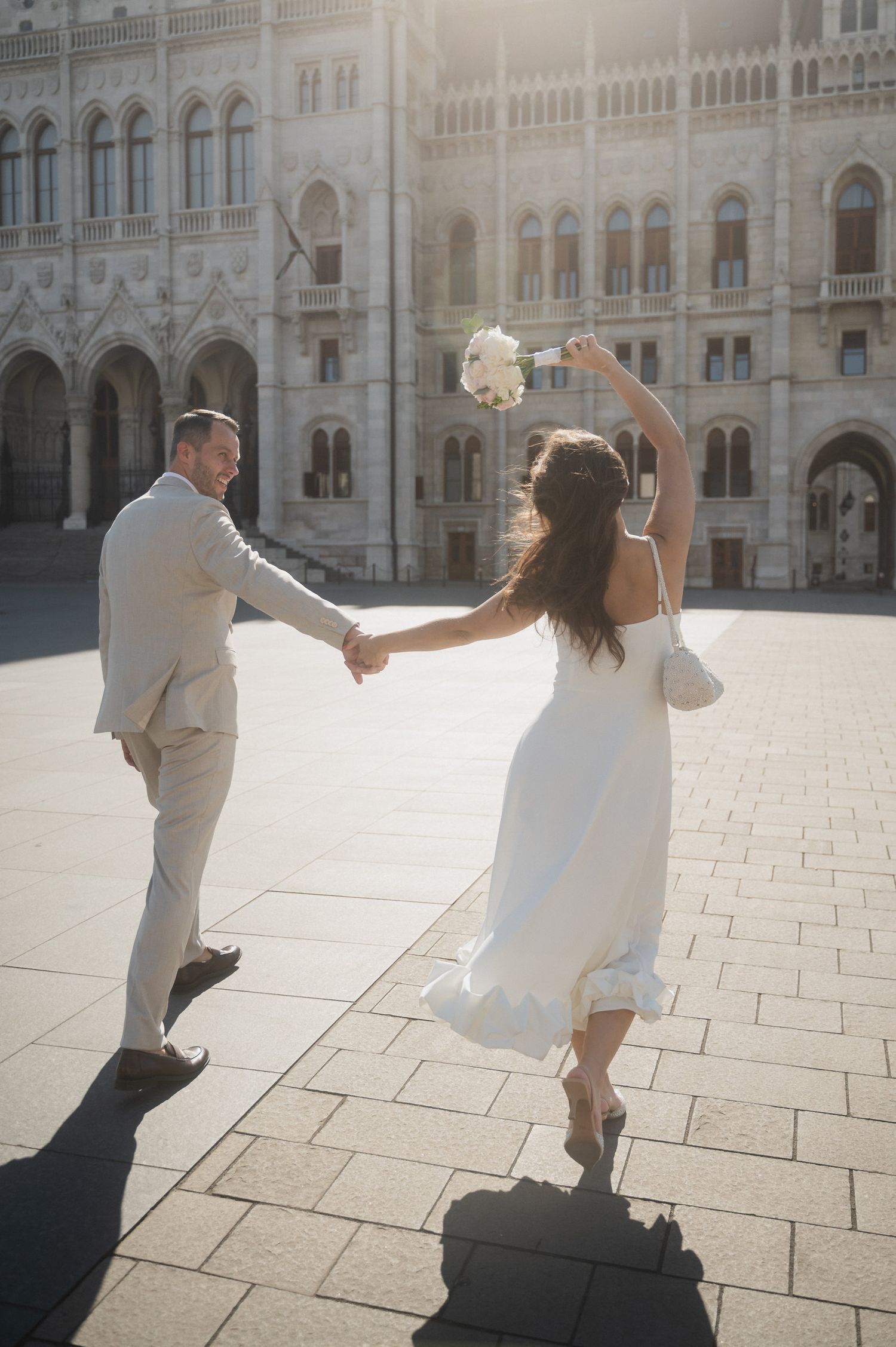 A couple in wedding attire dances joyfully in front of an ornate European building.