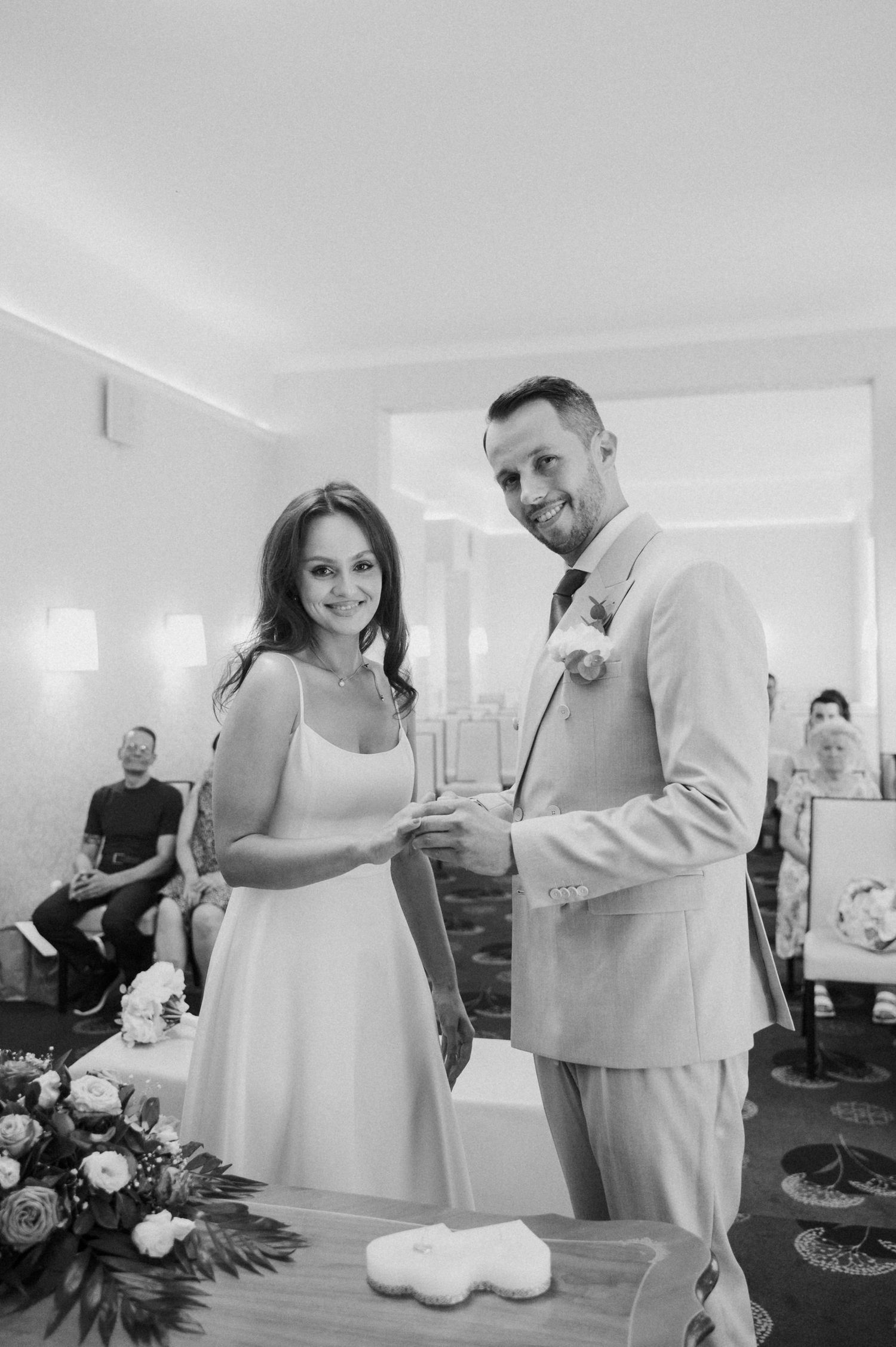 A couple in white and beige attire share an intimate moment while looking at something together indoors.