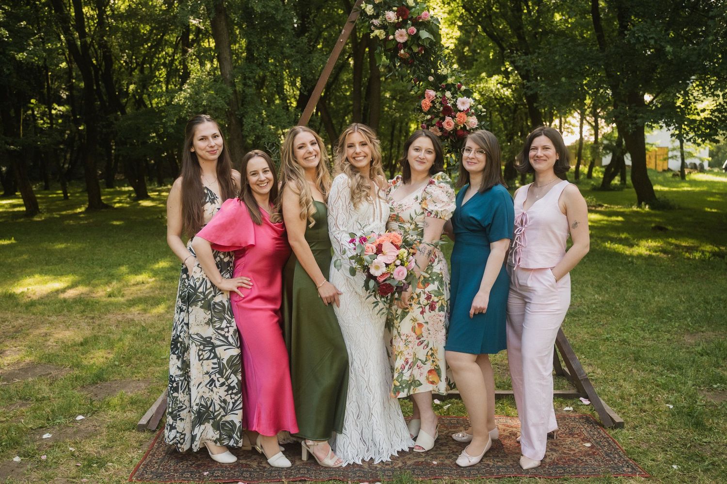 A group of people in colorful dresses and formal attire pose together for photos in a sunny outdoor garden setting.