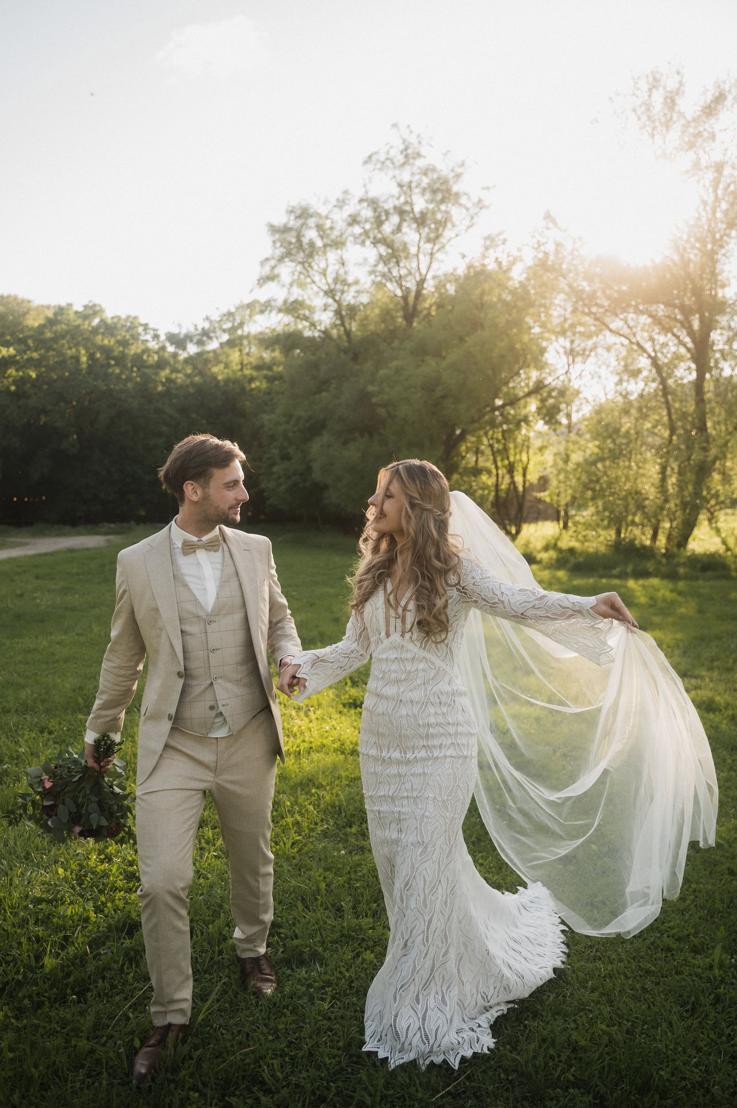 A couple in wedding attire dances and twirls in a sunlit meadow during golden hour, with the bride's veil flowing in the breeze.
