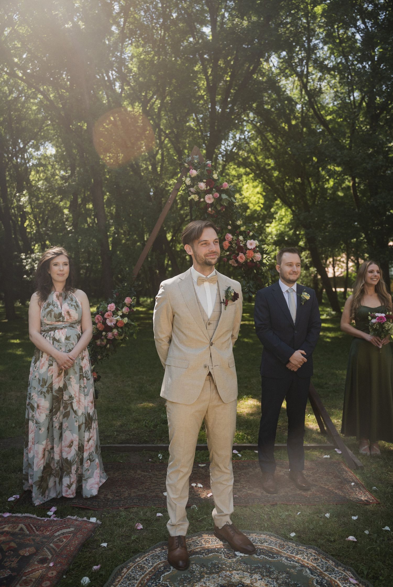 A wedding ceremony in a sunlit garden with attendants standing on a decorative rug surrounded by trees and floral decor.