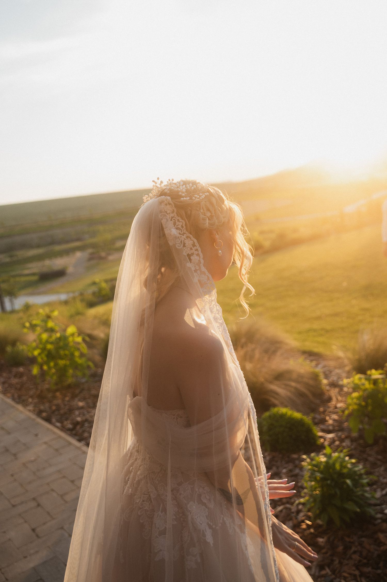 Bride in flowing veil silhouetted against golden sunset landscape.