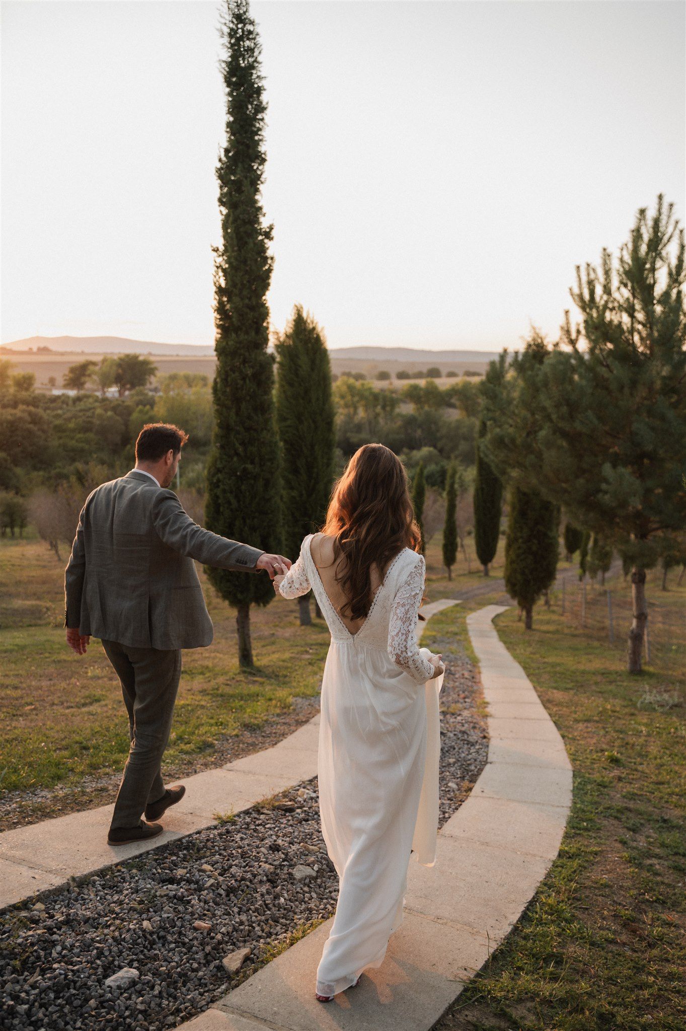 A couple walks along a winding path through cypress trees at sunset in a romantic outdoor setting.