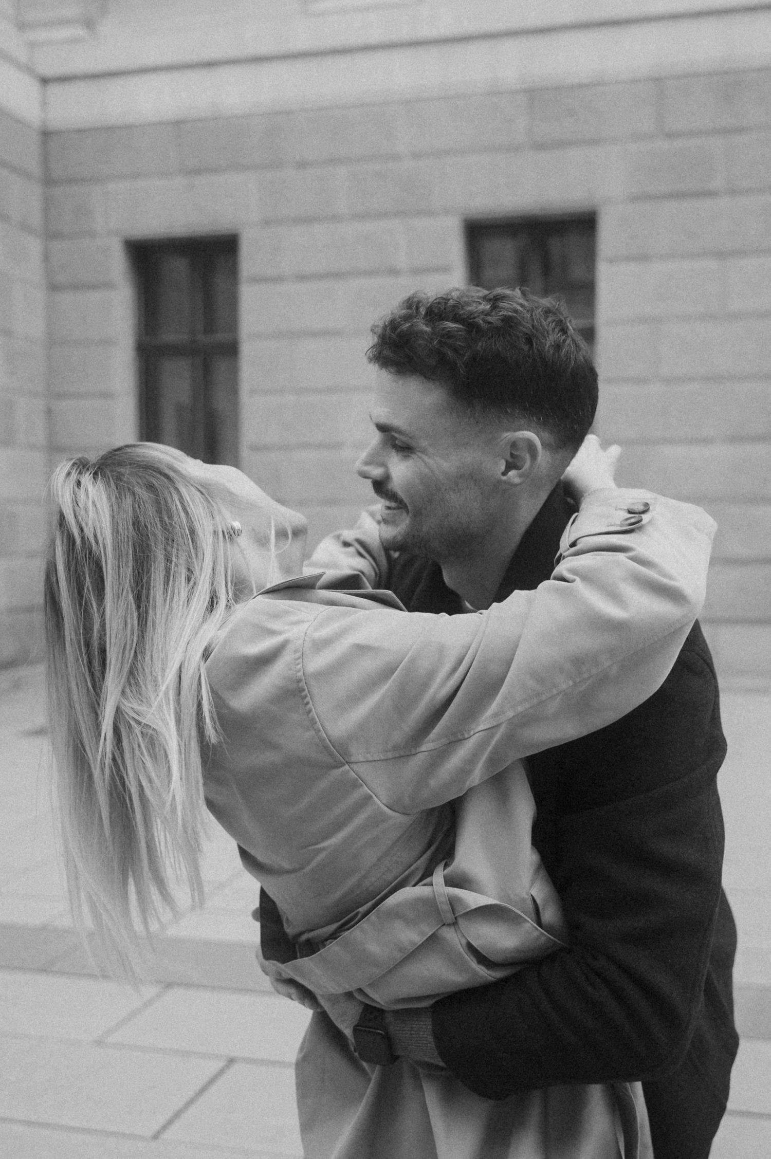 A romantic black and white photograph shows a couple embracing outdoors against a stone building wall.