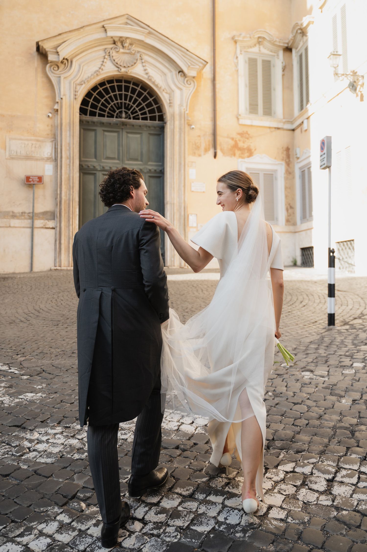 Newlyweds walk together on historic cobblestones with elegant architecture in the background.