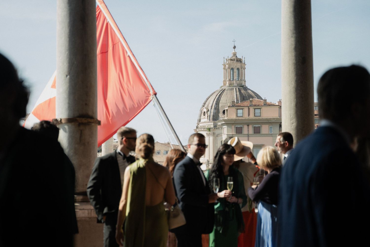 Red flags wave against a stone colonnade during an outdoor wedding celebration.