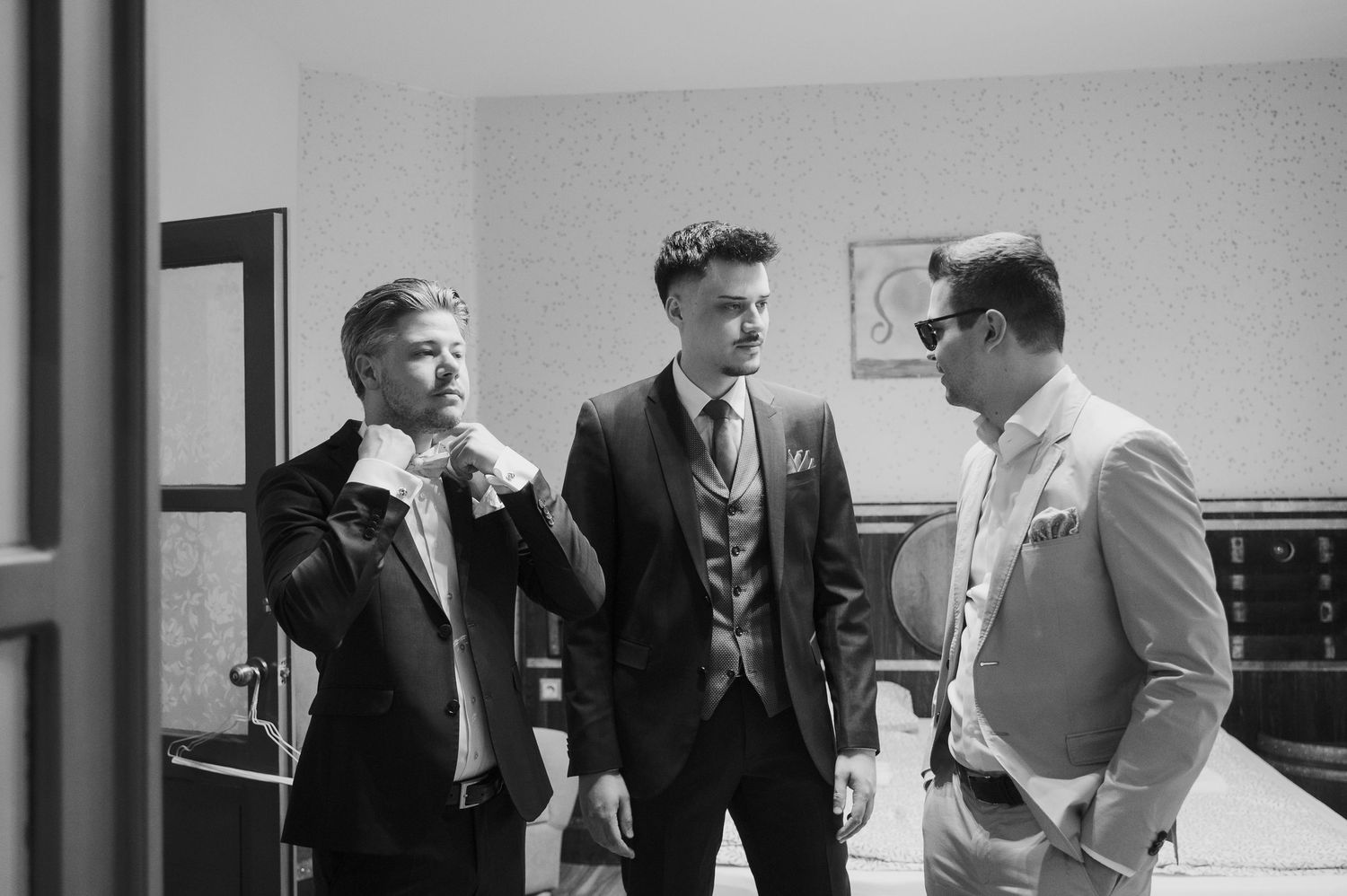 Three men in formal suits chat and adjust ties in a black and white wedding preparation photo.