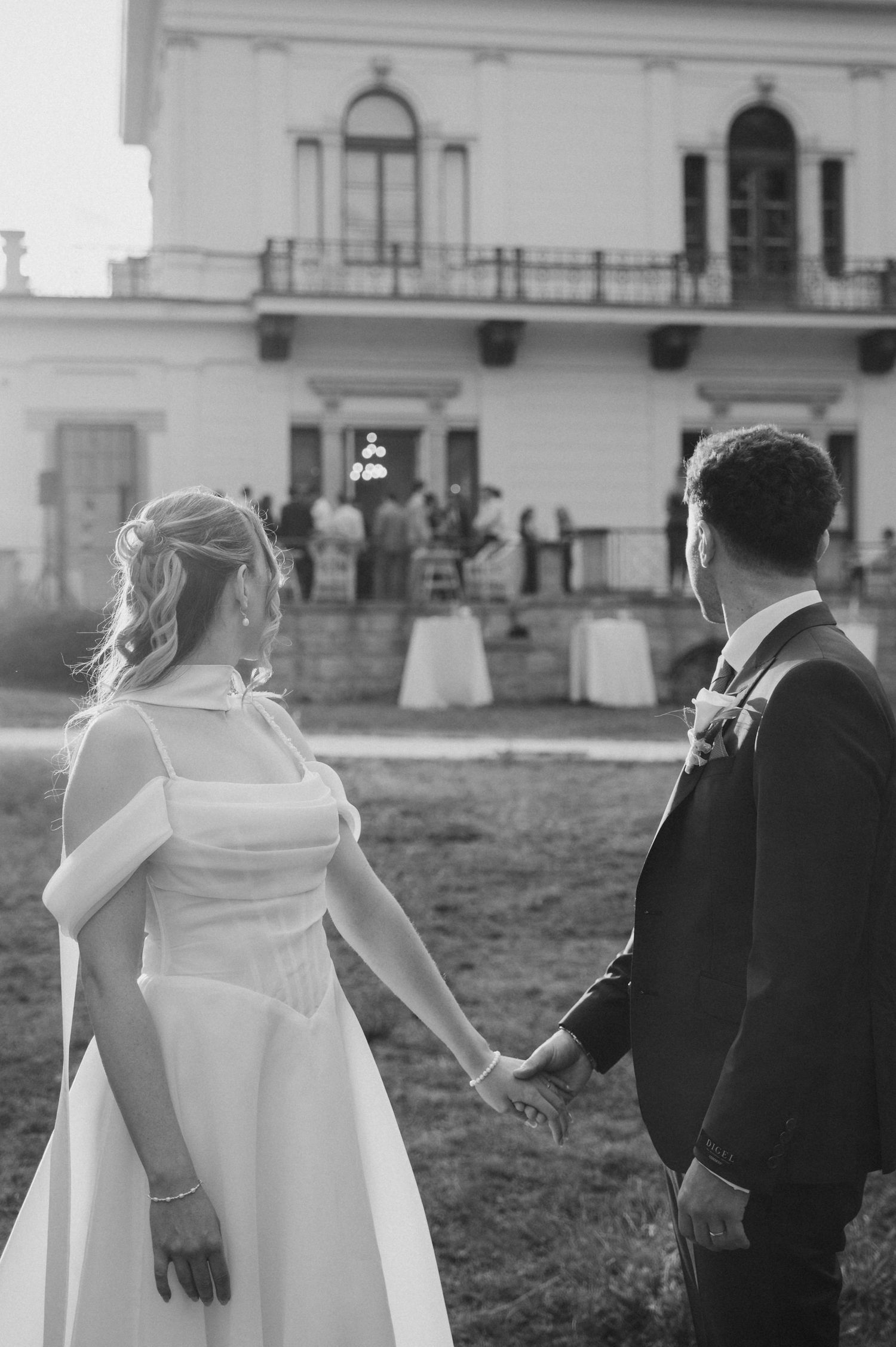Black and white photo of bride and groom holding hands facing elegant white villa.