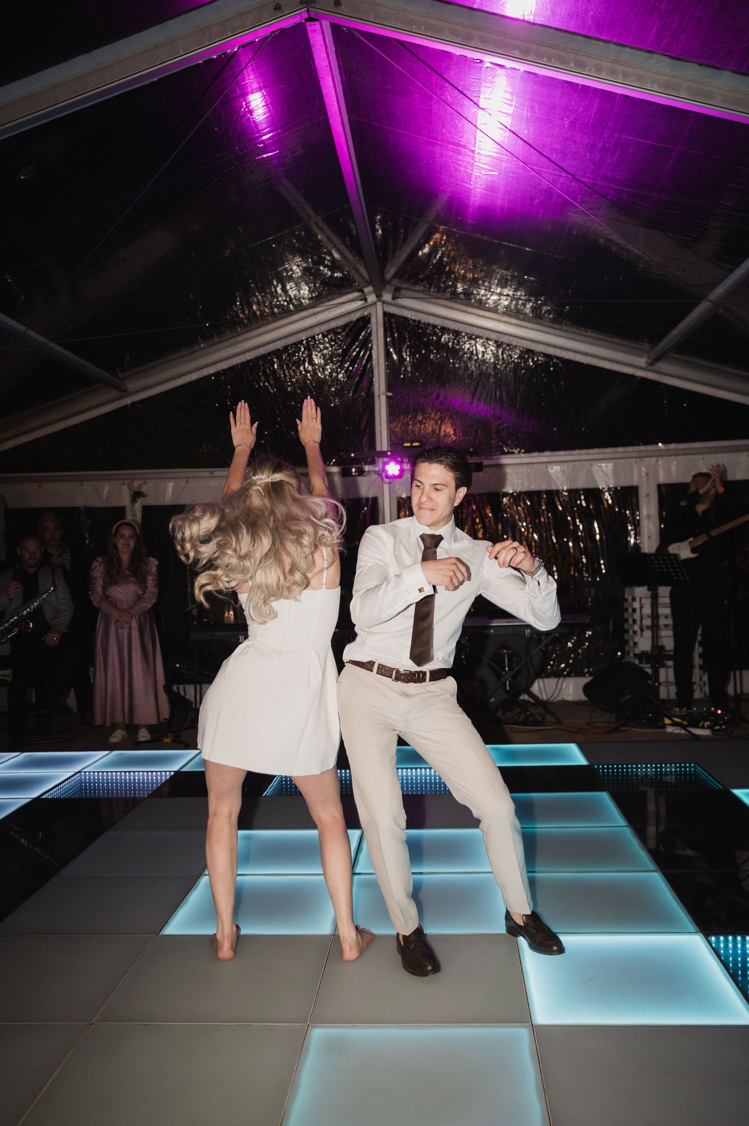 Bride and groom dancing energetically on illuminated LED dance floor under purple lights at reception.