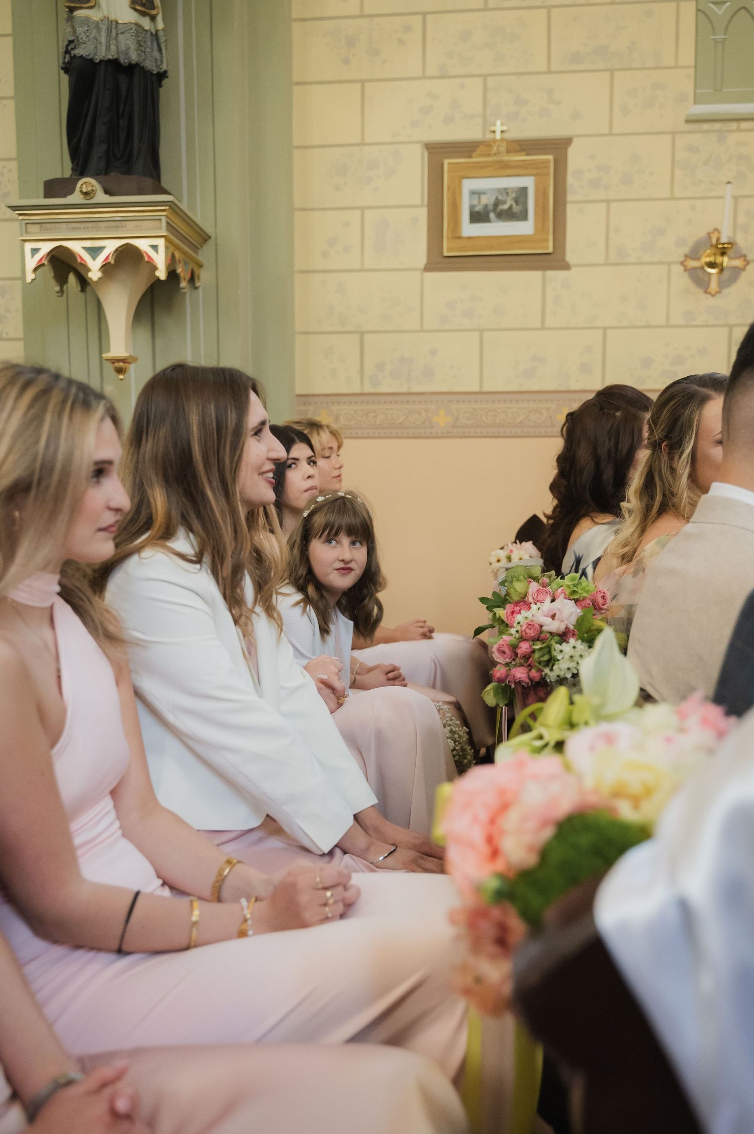 Wedding guests seated in a pastel church, smiling warmly during a ceremony with floral bouquets.