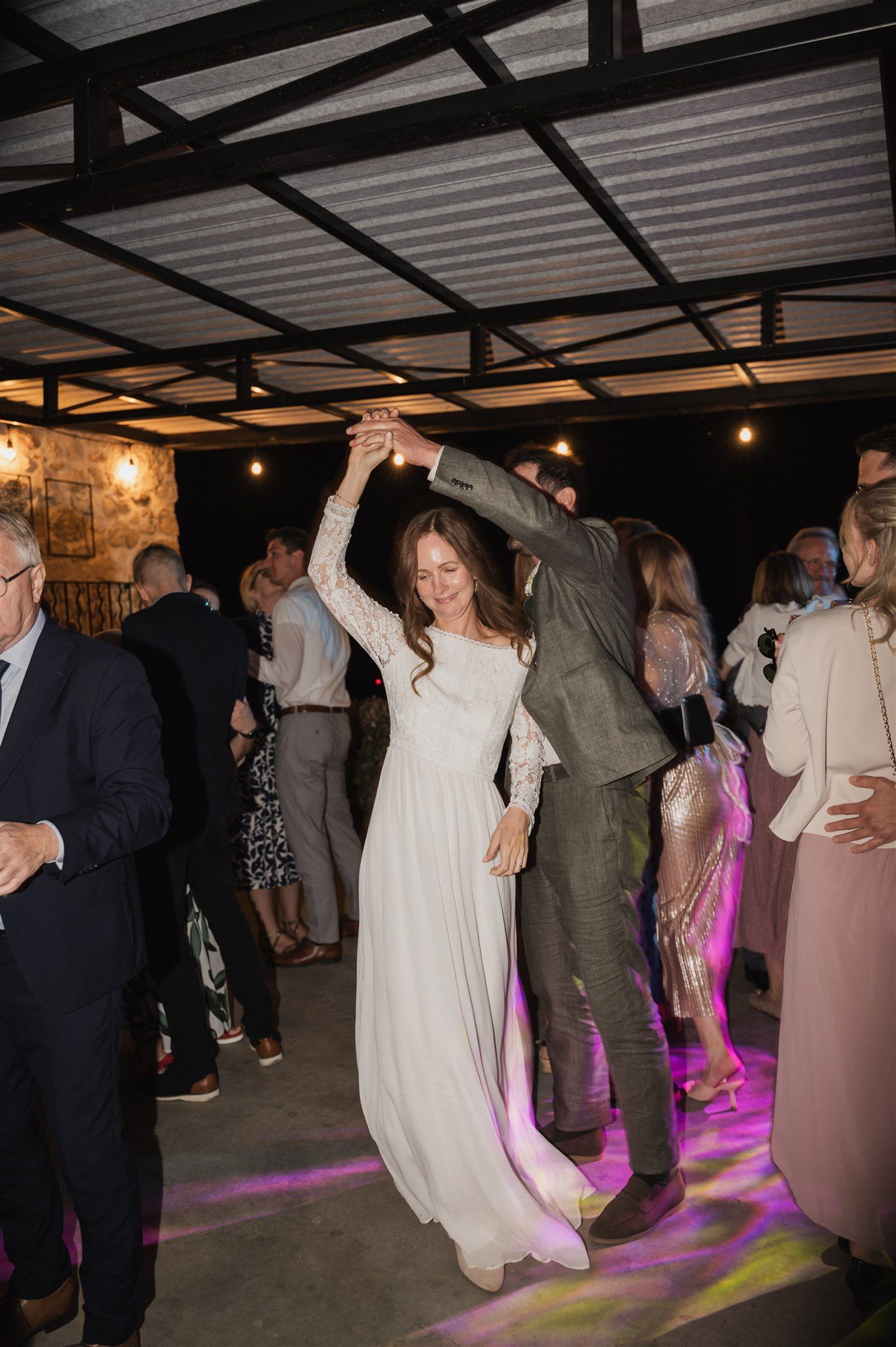 Wedding guests dance under string lights at an indoor reception with purple lighting effects.
