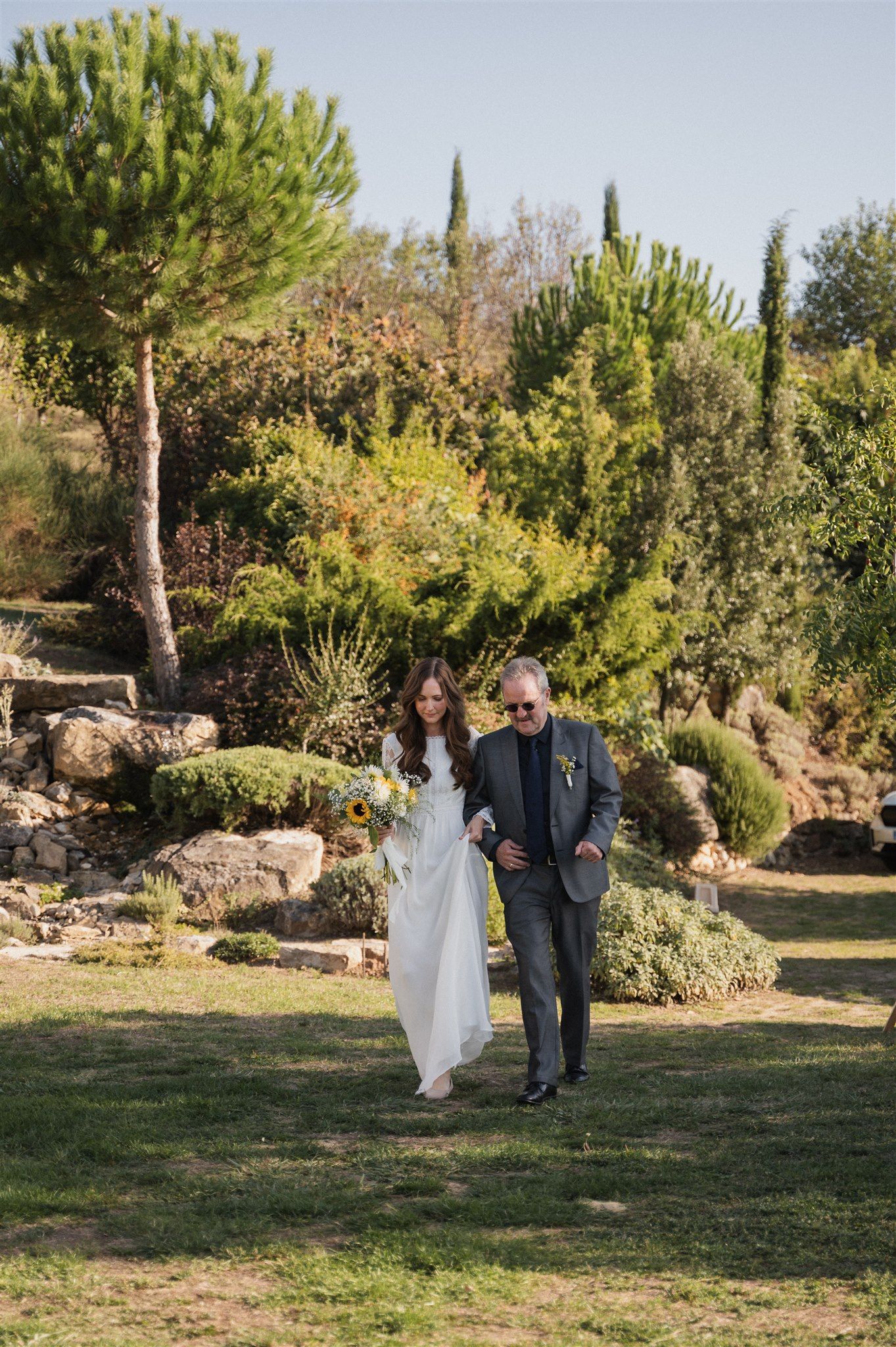 Couple walking together on grass with cypress trees and garden landscaping in background.