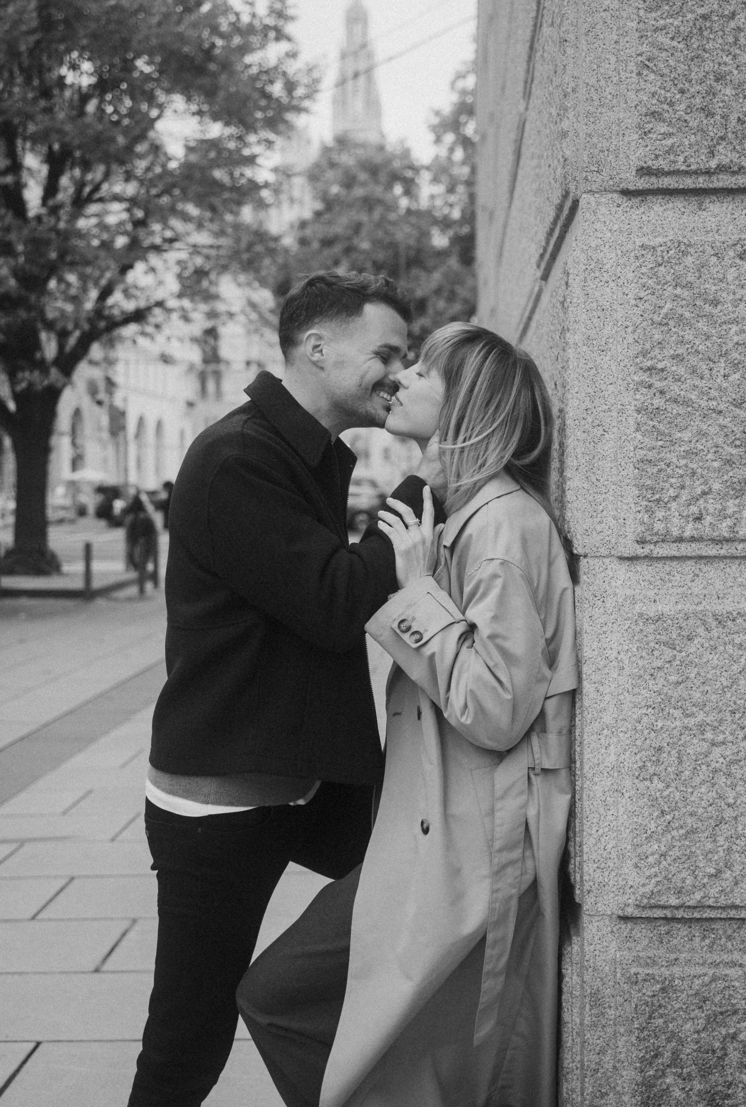 A romantic black and white photograph of a couple sharing an intimate moment against a stone wall on a city street.