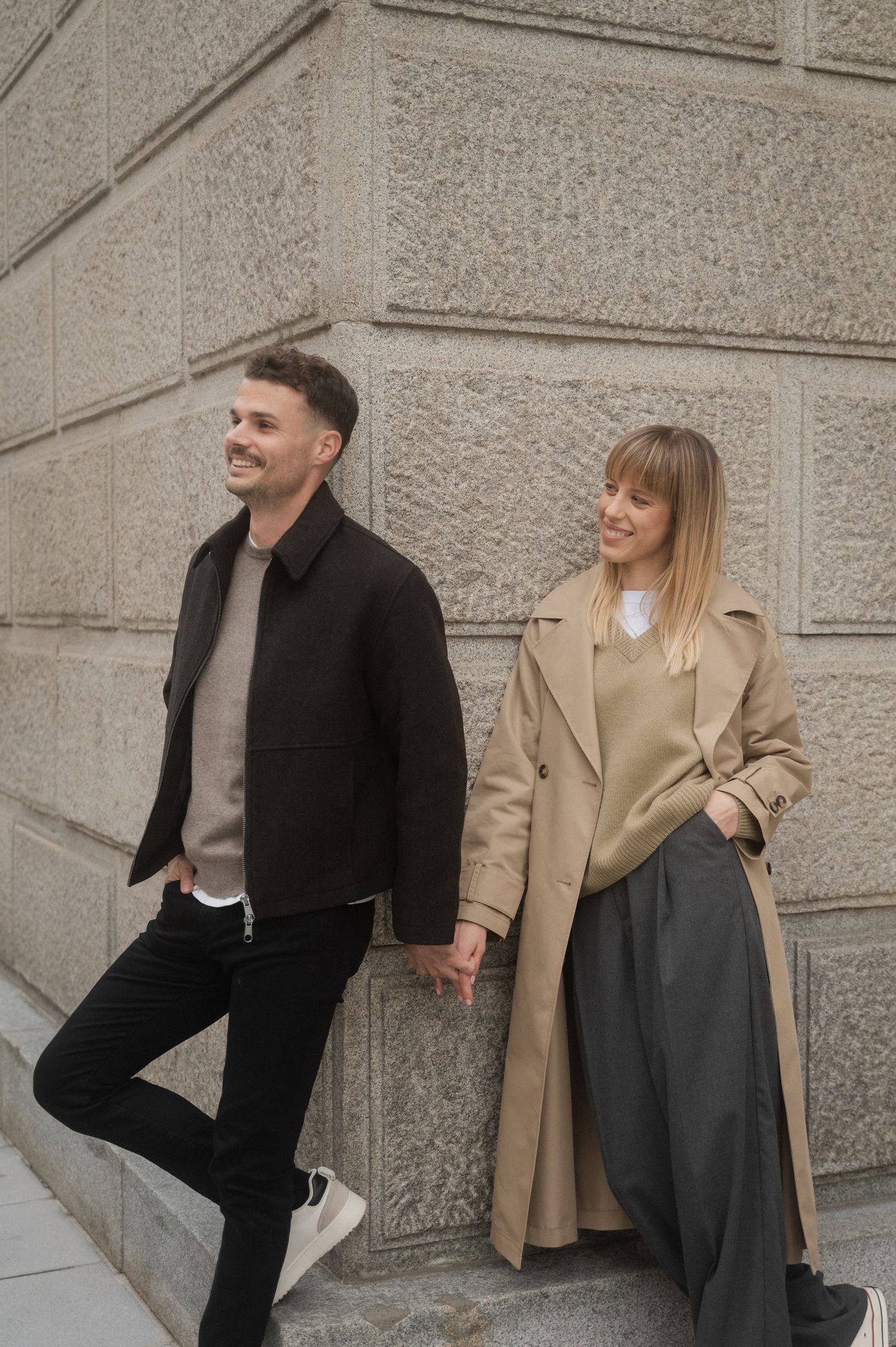 Stylish couple in winter coats lean against stone wall wearing casual urban outfits in neutral tones.