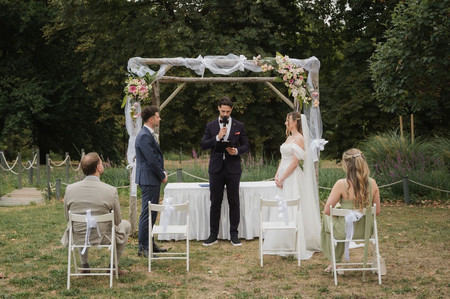 Outdoor wedding ceremony under floral arch with couple, officiant, and seated guests in lush garden setting.