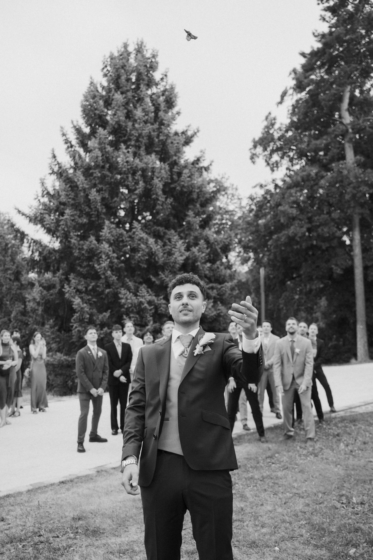 Groom in suit tossing bouquet outdoors with wedding guests gathered in background, black and white photo.