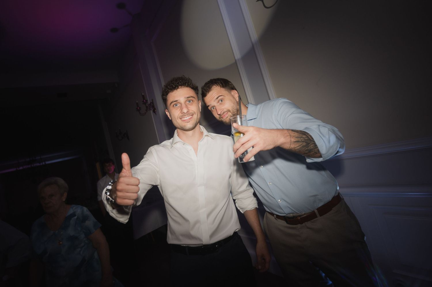Two smiling male wedding guests pose together with thumbs up and drinks at reception party.