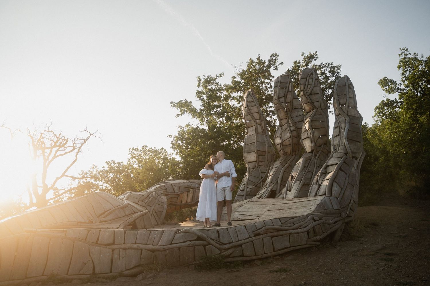 A couple in white outfits stands on a rocky cliff overlooking a sunset landscape.