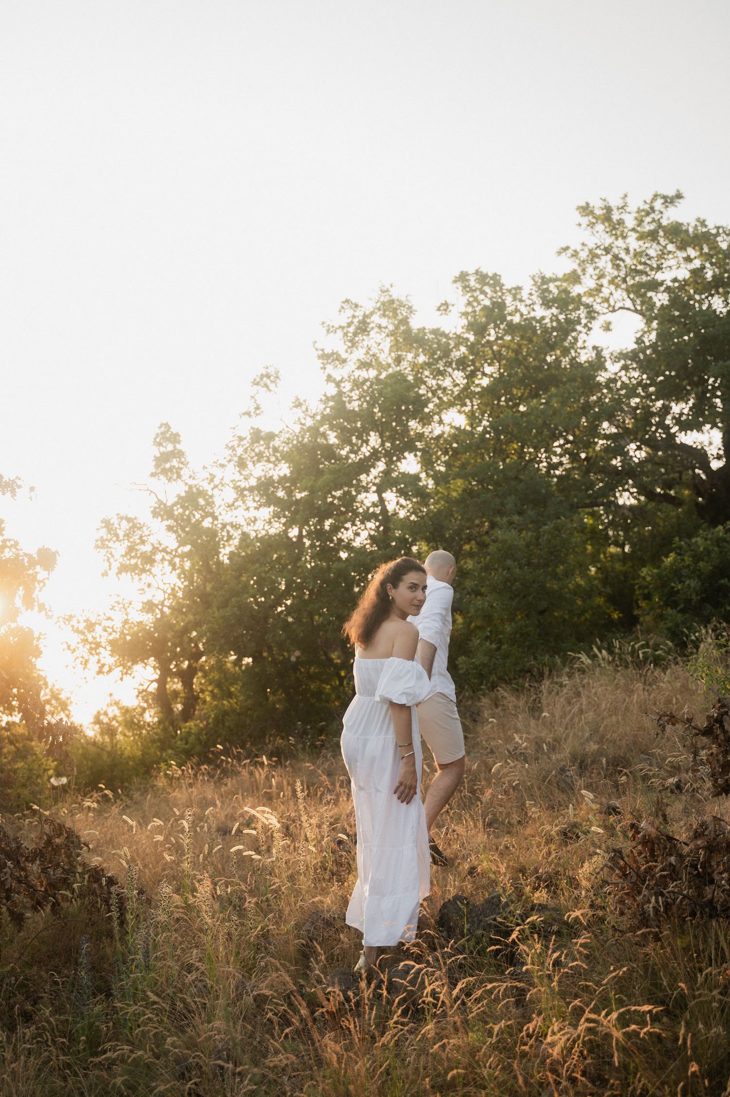 A figure in a flowing white dress stands in a sunlit field of tall golden grass at sunset.