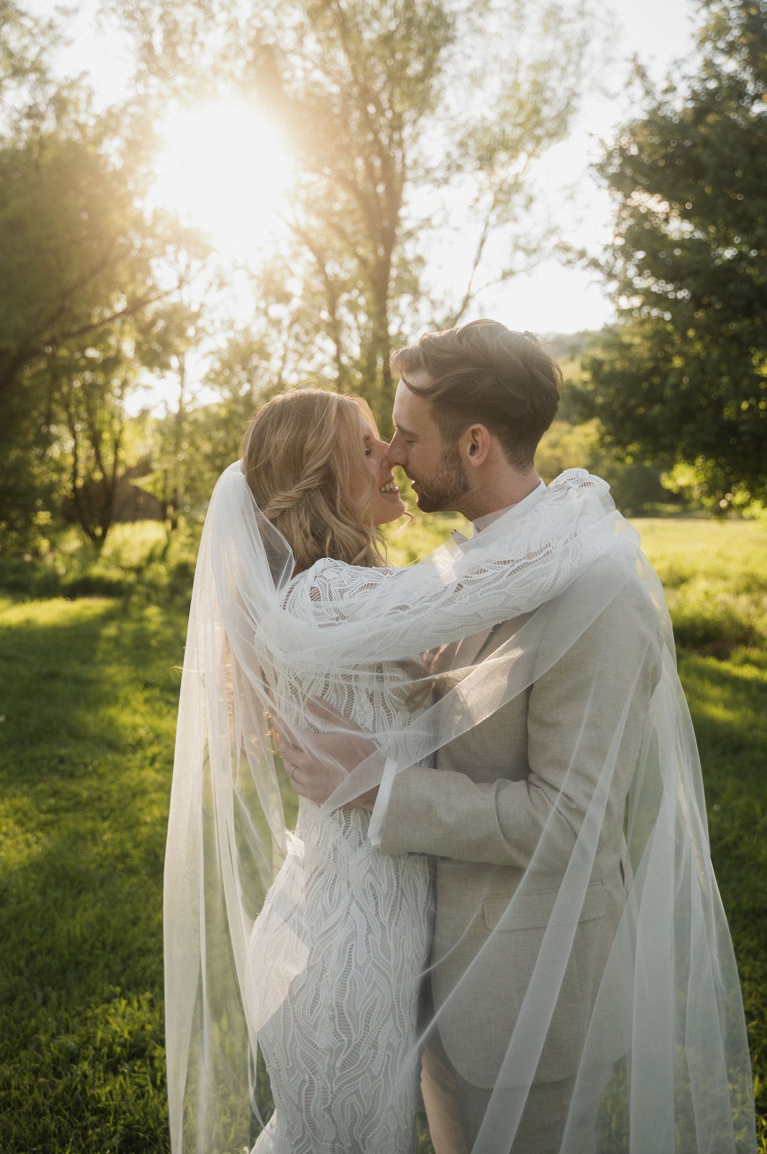 A romantic wedding couple embraces in a sunlit garden setting with flowing veil and natural backlight through trees.