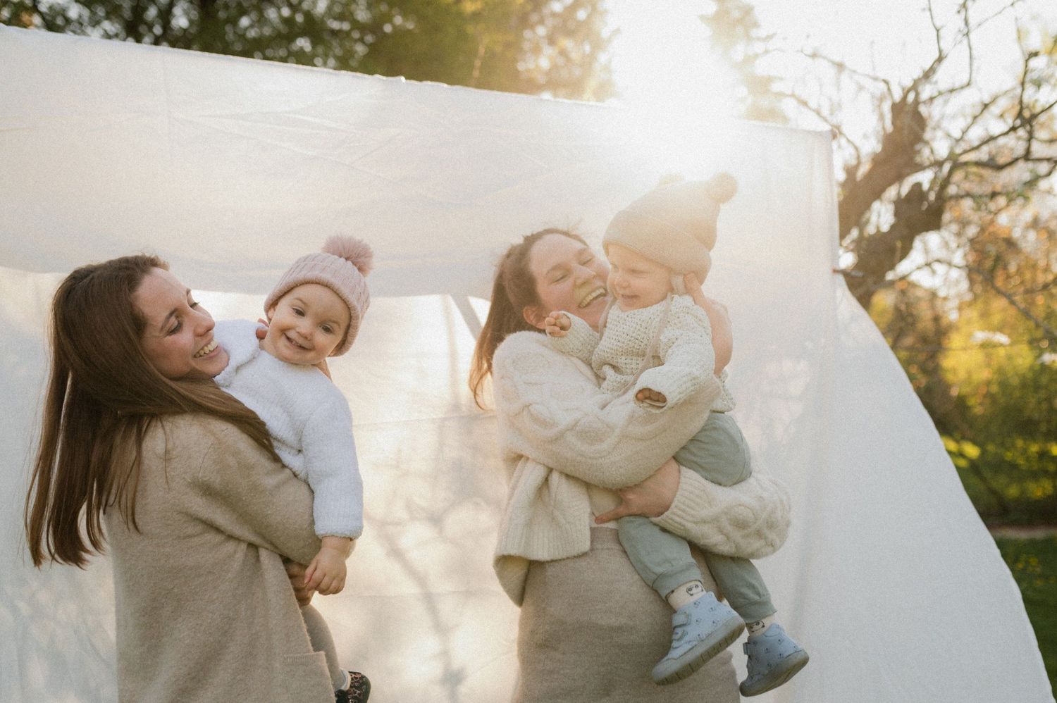 Two mothers laughing outdoors holding toddlers in soft sunlight with white fabric backdrop.