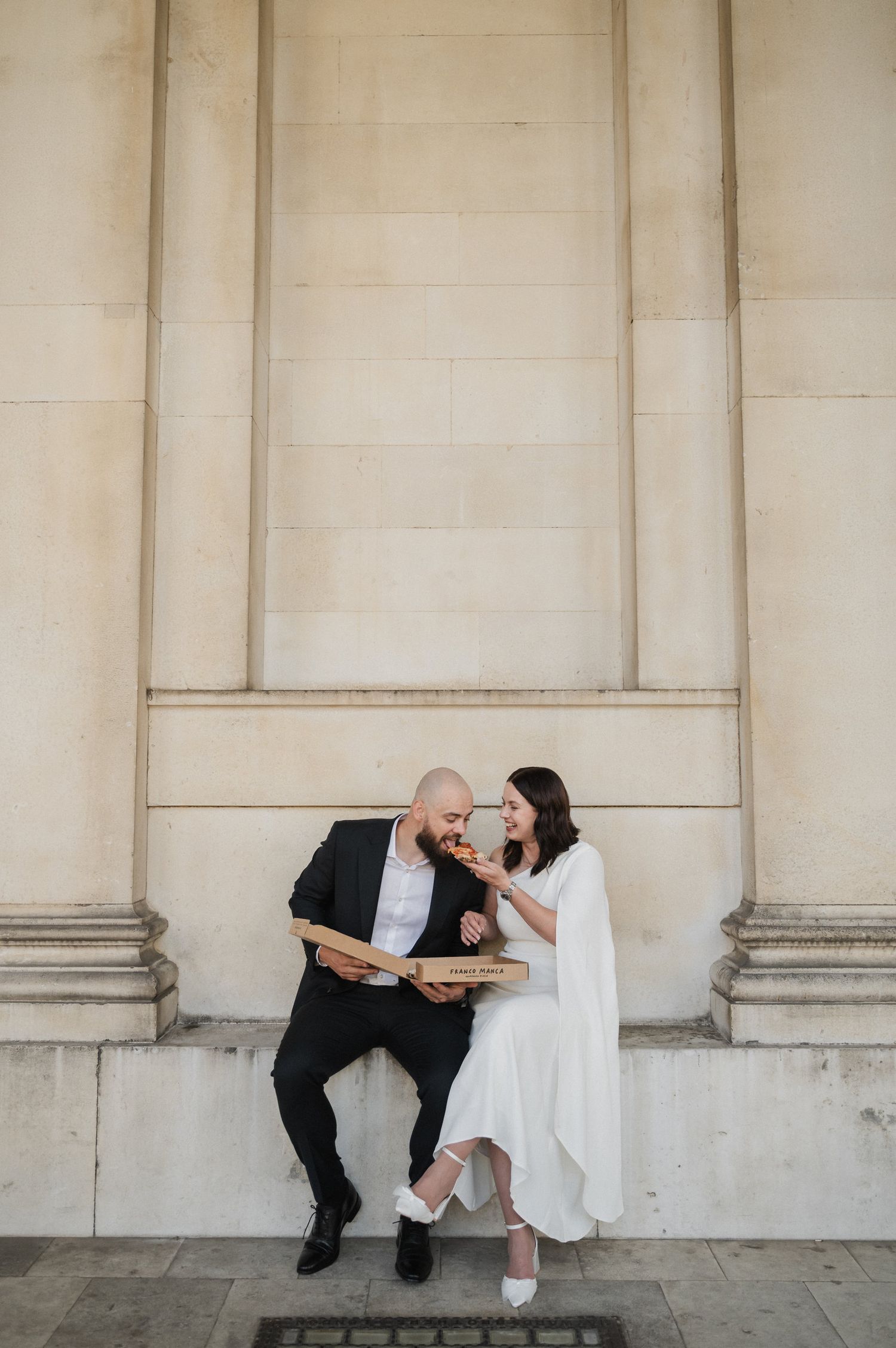 A playful couple in wedding attire shares a laugh against a grand stone wall backdrop.